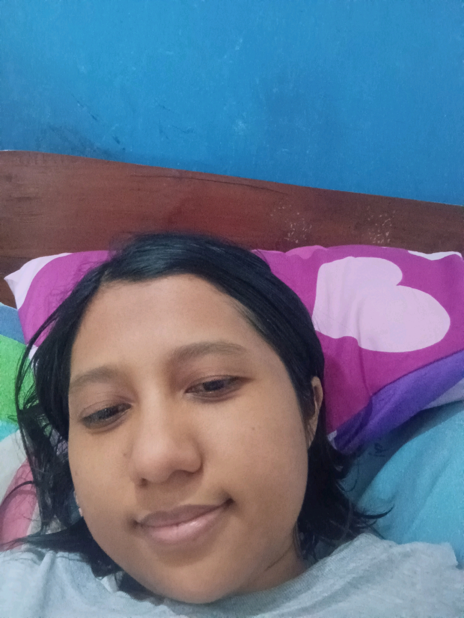 The image is a selfie taken by a young woman who appears to be lying down in bed. The background consists of a blue wall and a wooden headboard. The woman is resting her head on a pillow with a pink and white heart pattern. She has dark hair and is wearing a grey t-shirt. The lighting suggests the photo was taken indoors. The image does not provide any specific details about the city of Canga'an, Indonesia.