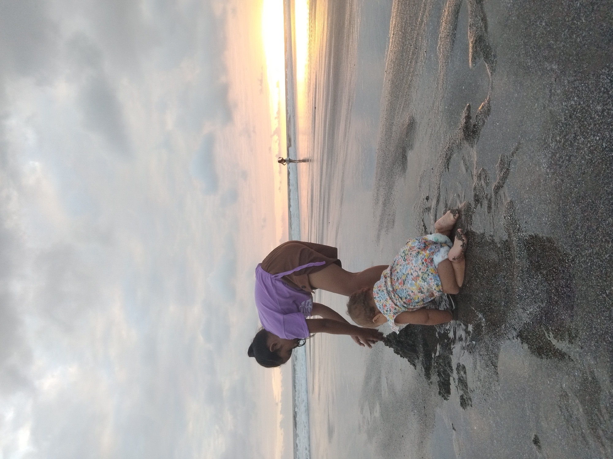 Two young females are on a beach in North Kuta, Indonesia. The older female, wearing a purple t-shirt and brown shorts, is bent over, playing with wet sand. A younger female, in a floral-patterned romper, is kneeling beside her, also interacting with the sand. The sun is setting, casting a warm glow on the horizon. The ocean waves are visible in the background, and a lone figure stands further down the beach. The sand is dark and wet, with some puddles reflecting the sky. The sky is filled with clouds, with the sun breaking through on the right side.