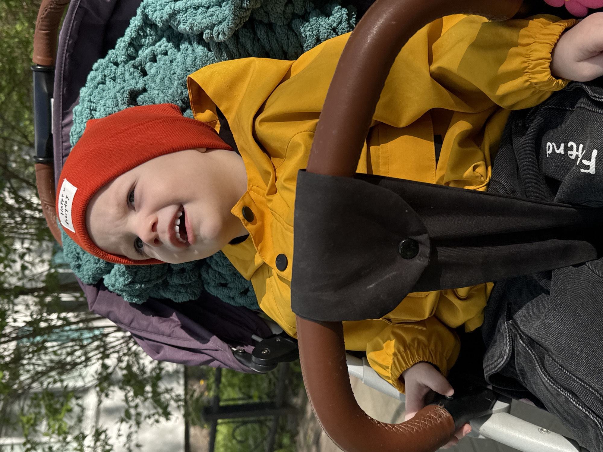 A toddler, wearing a red beanie and yellow jacket, is seated in a stroller. The stroller has a brown handle and a teal blanket inside. The child is wearing jeans with the word "Friend" embroidered on them. The photo was taken outdoors, with trees and a fence visible in the background. The location is potentially Saint Petersburg, Russia, given the context of the request.