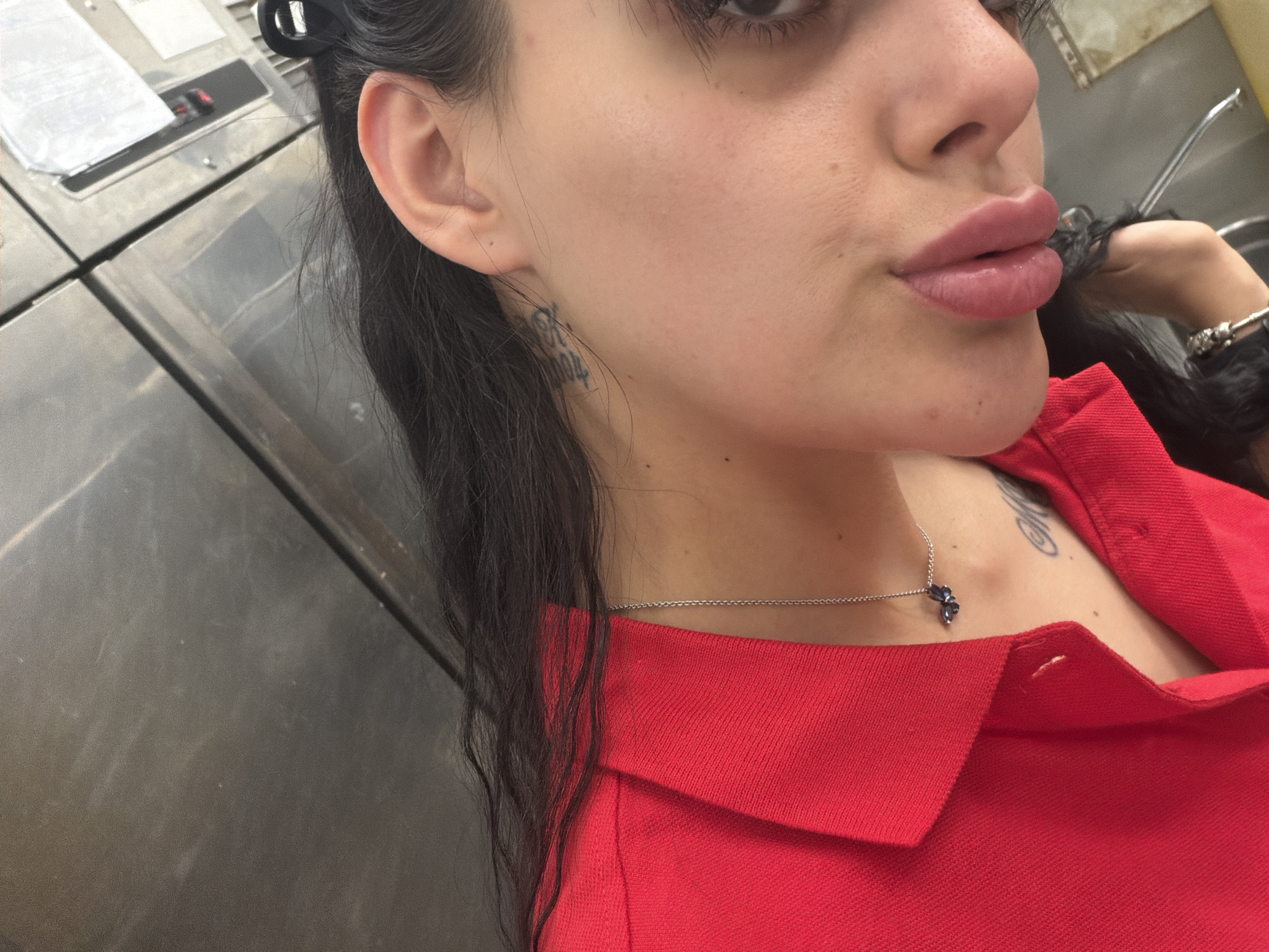This is a close-up, indoor, waist-up photograph of a person. The subject is a young woman with dark hair, wearing a red collared shirt. She has a small, dark tattoo on the side of her neck, and a delicate silver necklace with a blue butterfly pendant resting on her collarbone. Her lips are noticeably full and painted with a soft pink lipstick. Her left ear is visible, and she has a dark hair clip in her hair near her ear.
The background appears to be a metallic, industrial-looking surface, possibly part of a machine or structure, with some visible seams and a slight reflection. There is also a hint of a chrome faucet or pipe in the upper right corner. The lighting is artificial and appears to be from overhead, creating soft shadows. There is no indication of the time of day or weather conditions. No visible text is present in the image. The overall mood is casual and self-focused, suggesting a selfie. The setting might be a locker room, a laundry facility, or a similar utilitarian environment.
