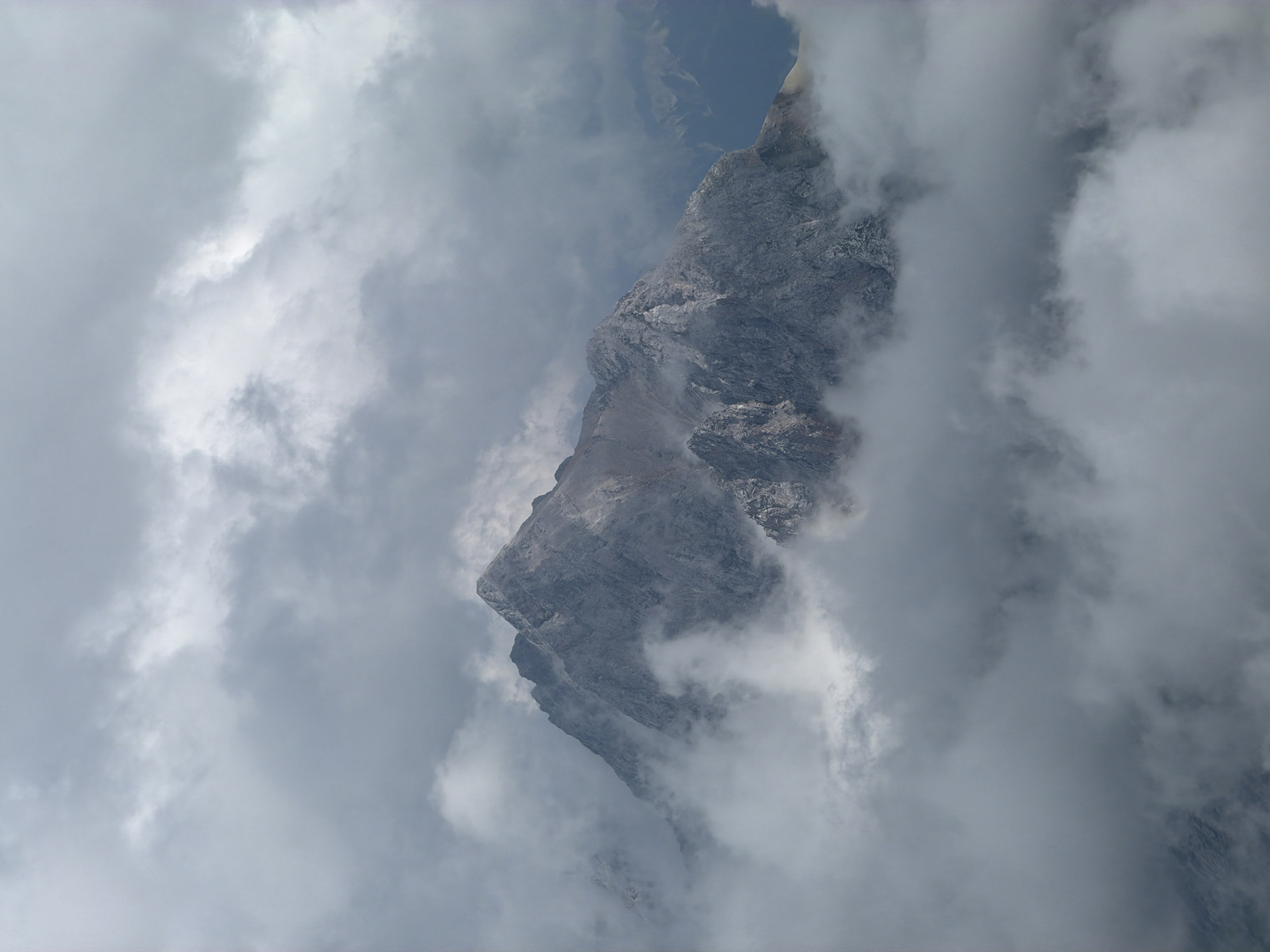 The image displays a dramatic mountain peak shrouded in clouds, characteristic of the Bavarian Alps. Specifically, this is likely Zugspitze, Germany's highest mountain, located near Garmisch-Partenkirchen. The mountain is rendered in shades of grey and white, with its rocky, rugged slopes partially obscured by wisps of cloud. The sky above is overcast with thick, heavy clouds, contributing to the moody and atmospheric depiction of the alpine environment. No people or man-made structures are visible in the frame, emphasizing the raw, natural grandeur of the mountain landscape.