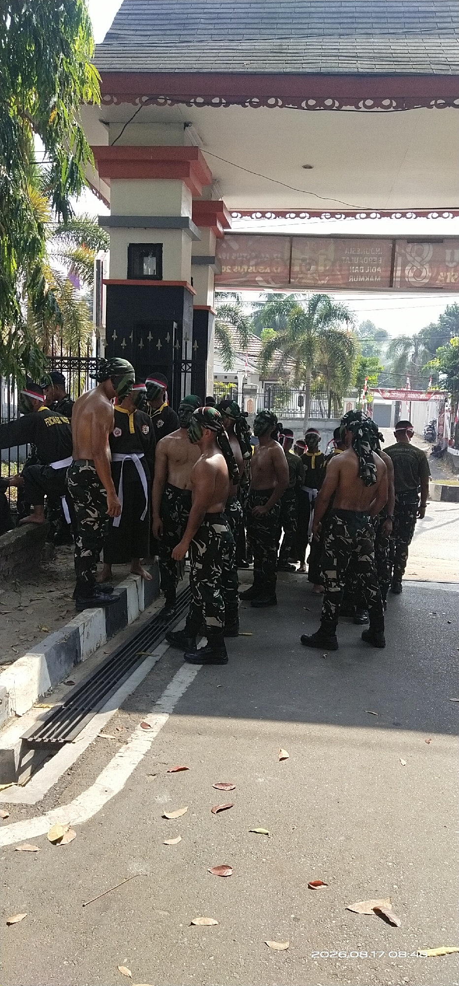In Tamiang Layang, Indonesia, a group of approximately 20 young men, appearing to be of Indonesian nationality, are gathered outside a building entrance. They are predominantly shirtless and wearing camouflage-patterned trousers. Several individuals have camouflage fabric wrapped around their heads, with some also having face paint in green and black. Two individuals on the left are wearing black uniforms with white sashes. The men are standing on a paved road next to a concrete curb with a metal grate drain. Behind them, a building with a tiled roof and pillars is visible. Palm trees and other vegetation are present in the background. The scene is illuminated by bright daylight, casting shadows on the ground. The timestamp on the image indicates it was taken on August 17, 2026, at 08:43.