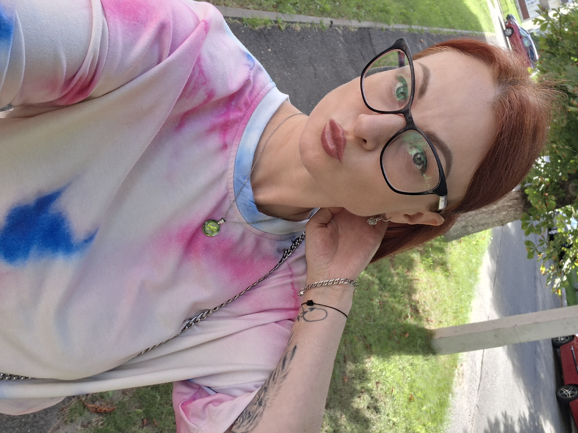 A woman with red hair and glasses is taking a selfie outdoors. She is wearing a tie-dye t-shirt with pink and blue colors. Her left hand is touching her neck, and she is wearing a bracelet and a necklace with a green pendant. There is a tattoo on her right arm. In the background, there is a tree, a sidewalk, grass, and a red car parked on a street. This scene is set in Myadzyel, Belarus.