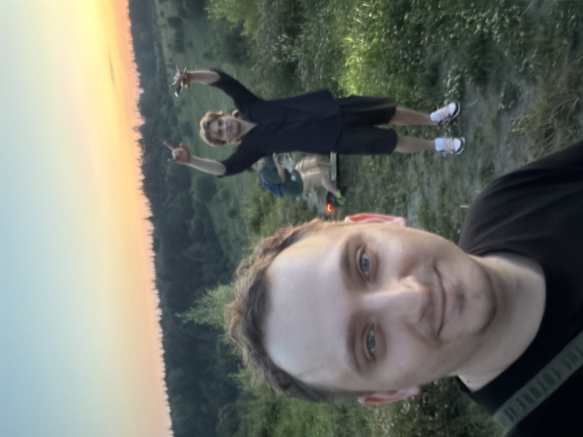 In this medium shot, a person's face dominates the foreground, with their head tilted towards the camera. They have light skin, blue eyes, and brown, curly hair. They are wearing a black t-shirt. The background appears to be a natural setting, with lush green foliage and a faint orange glow in the sky, suggesting either sunrise or sunset.
Further in the background, another person is visible, also facing the camera. This individual has blonde hair and is wearing a black t-shirt and dark shorts. They are striking a pose with their arms raised, and their legs are bent, indicating movement or a playful stance. They are wearing white and black sneakers. Behind this person, there are hints of other individuals and possibly a campsite with a small fire. The overall scene evokes a casual outdoor gathering in a rural or natural environment. The time of day is either dawn or dusk, given the warm hues in the sky.