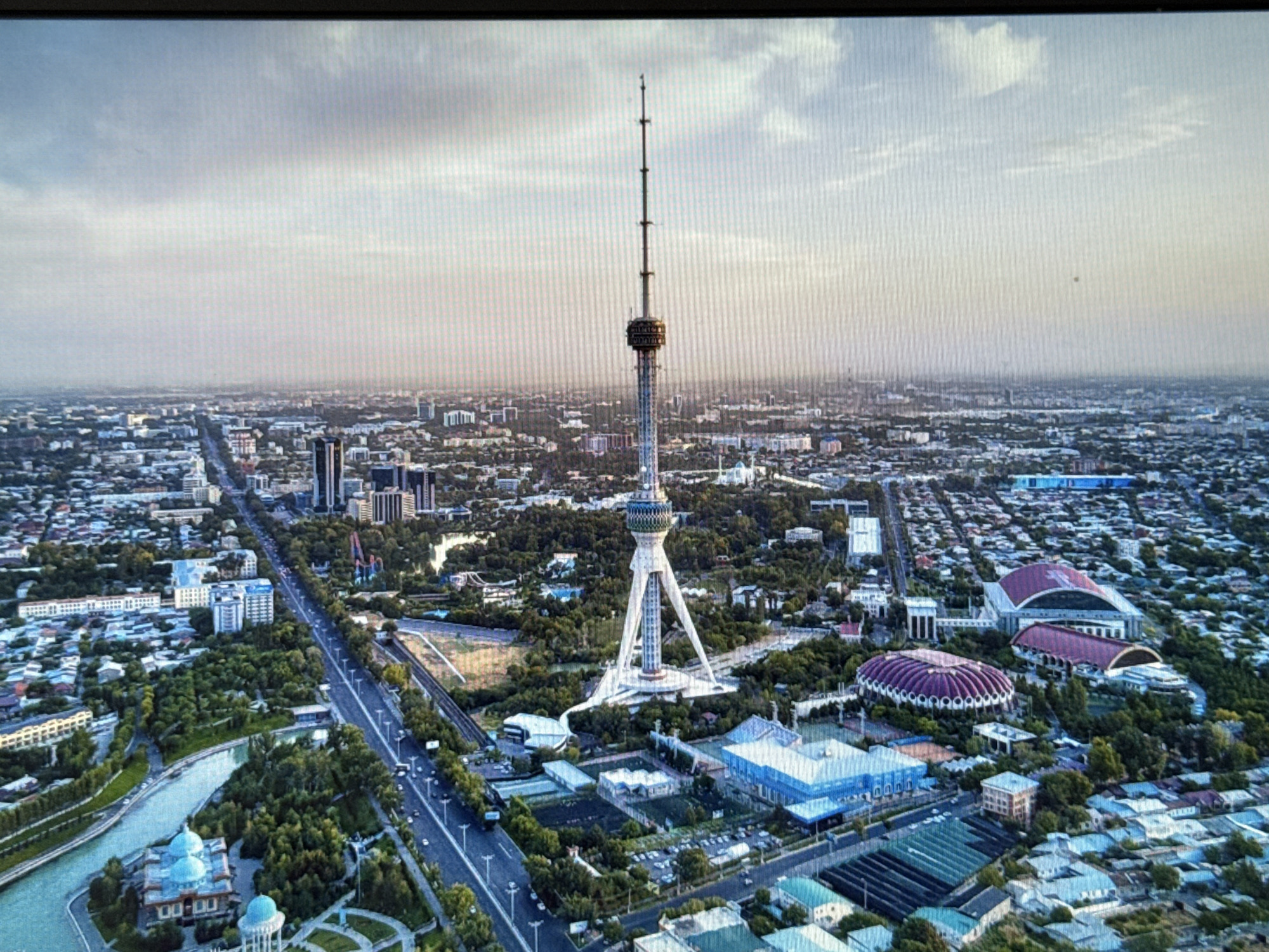 This is an aerial photograph of a city skyline dominated by a tall, slender television tower with a distinctive tripod base. The tower, which is the central focus of the image, pierces the sky, extending high above the surrounding urban landscape. The city itself is densely packed with buildings of varying heights and architectural styles, interspersed with green spaces, including trees and what appears to be a river or canal.
The scene is set during the daytime, under a cloudy sky that diffuses the light, creating a somewhat hazy atmosphere. The overall impression is of a sprawling metropolis, possibly in a warm climate, given the lush greenery.
Several notable structures are visible around the base of the tower, including modern buildings with blue roofs and other more traditional architectural designs with domed roofs. Roads with traffic can be seen winding through the city, indicating a bustling urban environment. In the middle ground, there are two large, distinctively shaped buildings, one with a red-tiled, curved roof and another with a prominent purple, ribbed dome.
While no specific activities or events are clearly depicted, the presence of traffic and the general urban layout suggest normal city life is in progress. The perspective is from a high vantage point, looking down and across the city, providing a panoramic view. No people are discernible in the image.
There is no visible text in the image. The location context provided (Pushkino, Russia) does not appear to be directly supported by any visual cues within the image itself, which shows a generic large city with a prominent TV tower.