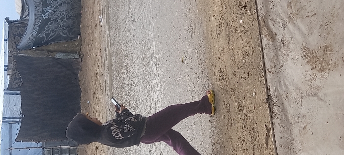 A child, seen in profile walking from left to right, is the central subject. The child wears a dark hooded sweatshirt with white lettering on the front, dark purple sweatpants, and bright yellow slip-on clogs. Both hands hold a light-colored smartphone horizontally, and the child's gaze is directed at the screen. The immediate ground consists of an uneven, light gray surface, potentially wet pavement or compacted dirt, leading to a drier, sandy area in the mid-ground. In the background, a series of makeshift shelters are visible, constructed from materials including light blue tarpaulins, dark fabric, and blankets, one featuring a prominent floral pattern. This scene is situated in the city of Khan Yunis, Palestinian Territory.