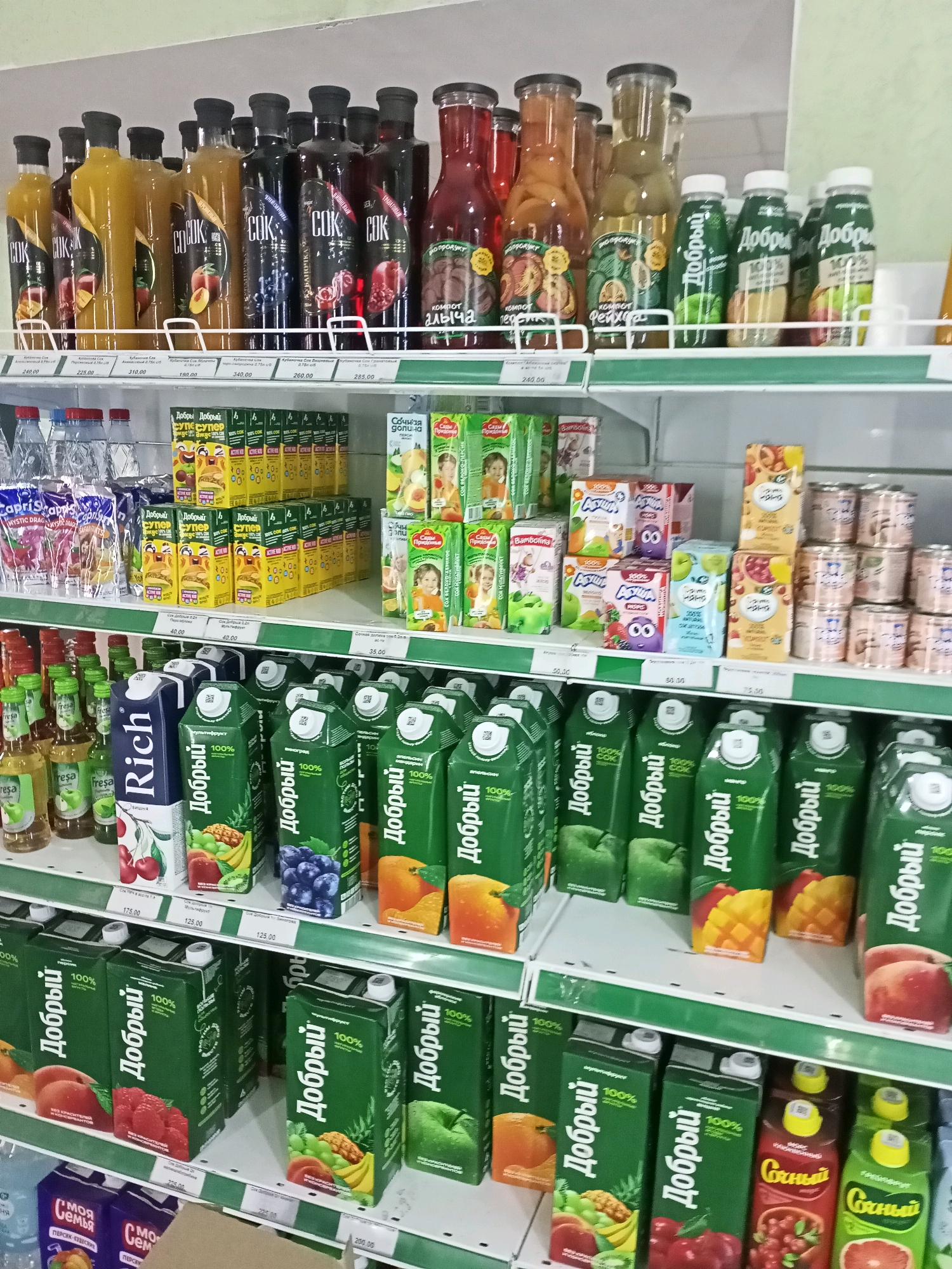 The image shows a grocery store refrigerator display case in Tsandrypsh, Abkhazia. The shelves are stocked with a variety of juices and beverages in bottles and cartons. The majority of the products are juices, with brands like "Добрый" (Dobry) and "Rich" visible. There are also some canned drinks and bottled water. The products are arranged in rows on multiple shelves. The lighting is bright, typical of a retail environment. There are no people visible in the image.