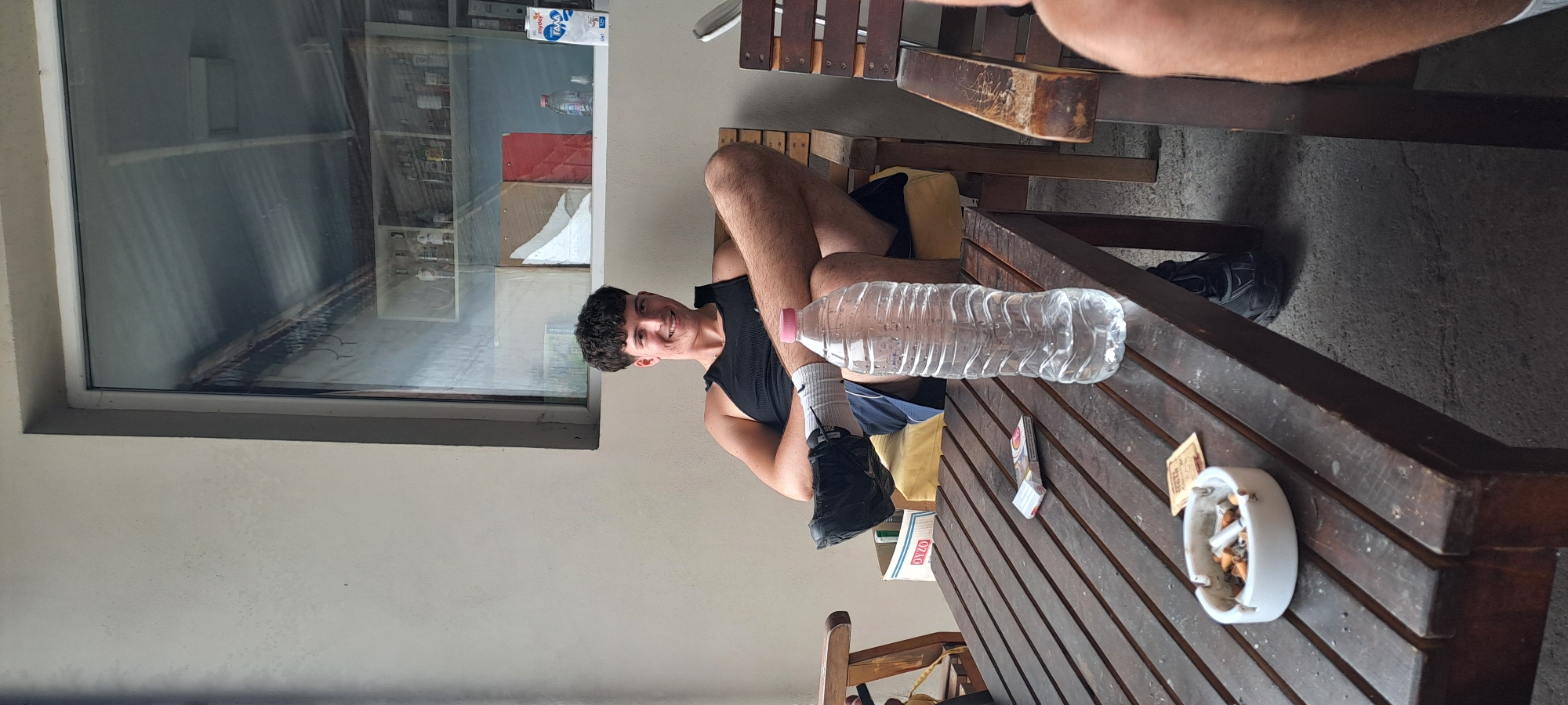 The image, rotated counter-clockwise, depicts a young man seated at a wooden outdoor table in what appears to be a cafe or patio setting in Velika, Bulgaria. The young man, positioned on the left side of the frame, has dark, curly hair and is wearing a black tank top and black shorts. He is smiling at the camera, conveying a relaxed and friendly demeanor. His legs are visible, with white socks featuring a "Nike" logo and black athletic shoes.
On the dark wooden table in front of him rests a large, clear plastic water bottle with a pink cap. To the right on the table, there is a white ceramic ashtray filled with several cigarette butts. Beside the ashtray, a small paper item is visible, possibly a receipt or wrapper, with some indistinct text that might include "Скоч" (Scotch). Another small box or packet, possibly labeled "ONZO," is also on the table near the man's legs.
A large window dominates the background to the left, reflecting an interior space with shelves, various items, and what seems to be an electrical panel. Through the window reflection, a white carton with blue text, possibly reading "mydos" and "ΓΑΛΑ" (Greek for milk), is discernible. The setting includes light-colored walls and a concrete ground, with parts of another person's arm or leg visible in the upper right, suggesting additional company. The bright, natural lighting indicates it is daytime.