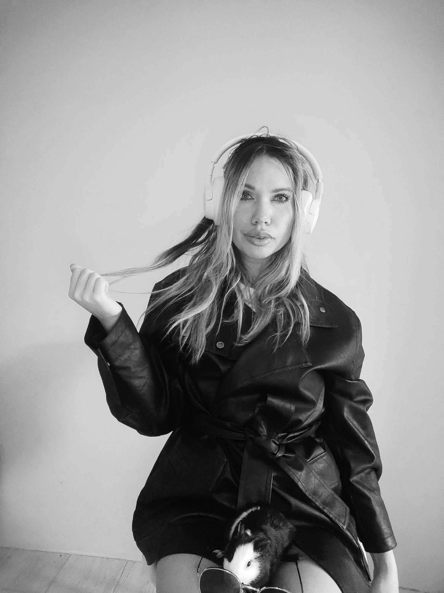 The black and white image shows a woman wearing white headphones and a black leather jacket, posing in front of a plain, light-colored wall. She is holding a strand of her hair in her left hand. On her lap is a guinea pig, with sunglasses placed near it. The image appears to be taken indoors. There is no information suggesting this image was taken in Votkinsk, Russia.