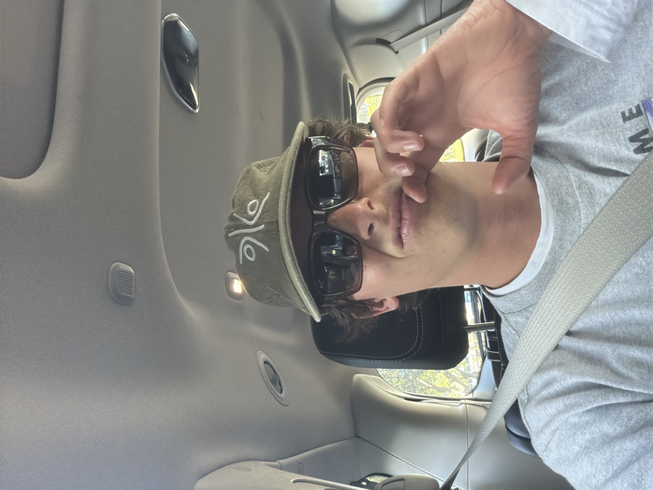 A person wearing sunglasses and a cap sits in a car, the seatbelt across their torso. The car's interior, including the ceiling with vents and lighting, is visible. The person's hand is near their face. A headrest is behind them. The image is rotated, as if taken with a phone and oriented sideways. The person appears to be making a gesture or posing for the camera.