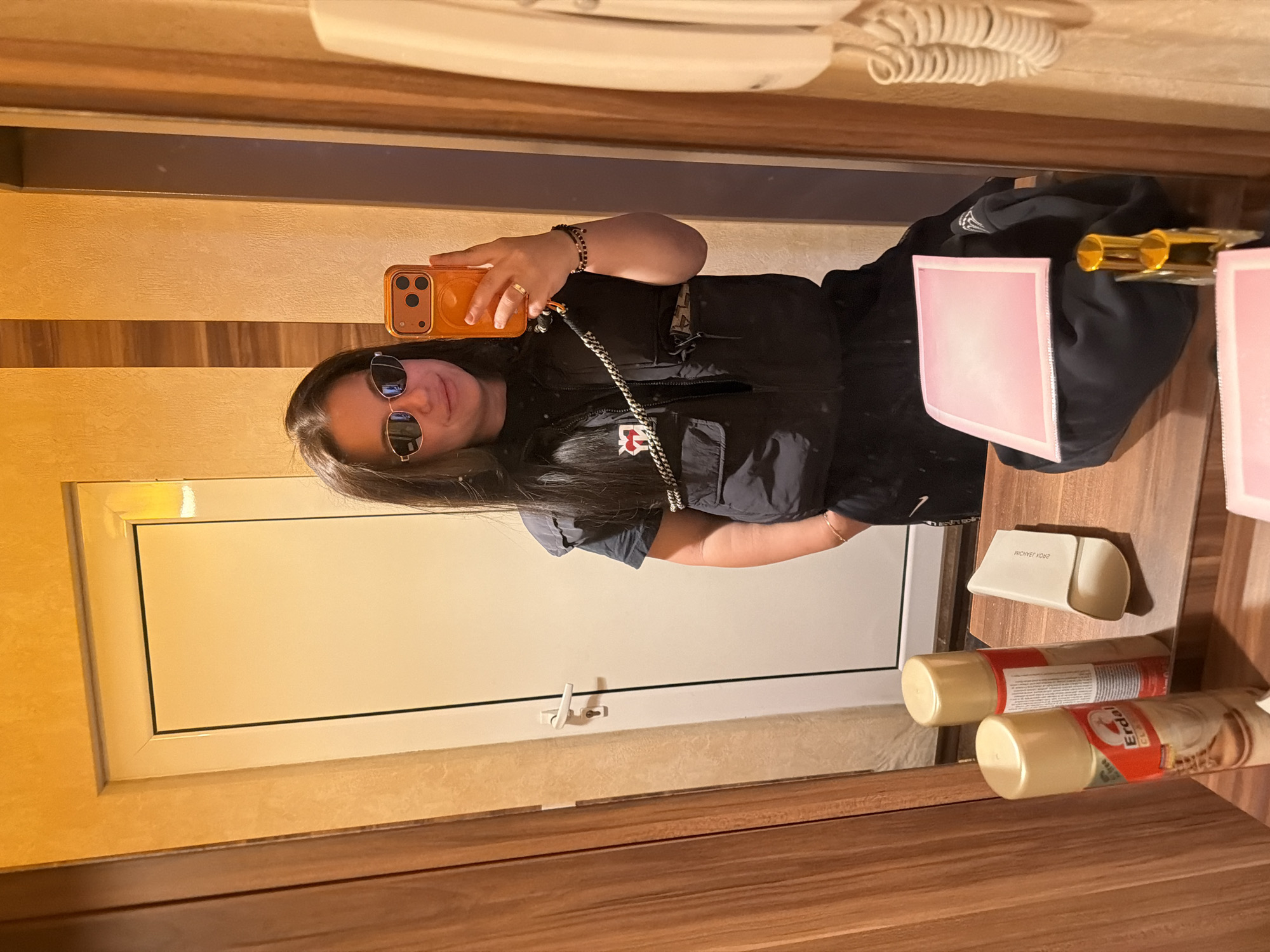 A woman is taking a selfie in front of a mirror. She has long dark hair and is wearing sunglasses and a black outfit. The mirror reflects a bathroom counter with cans of Erdal shoe polish and other items. The background shows a wooden door or wall panel. The location is likely a hotel or private residence. There is no explicit evidence linking the scene to the city of Burgas, Bulgaria.