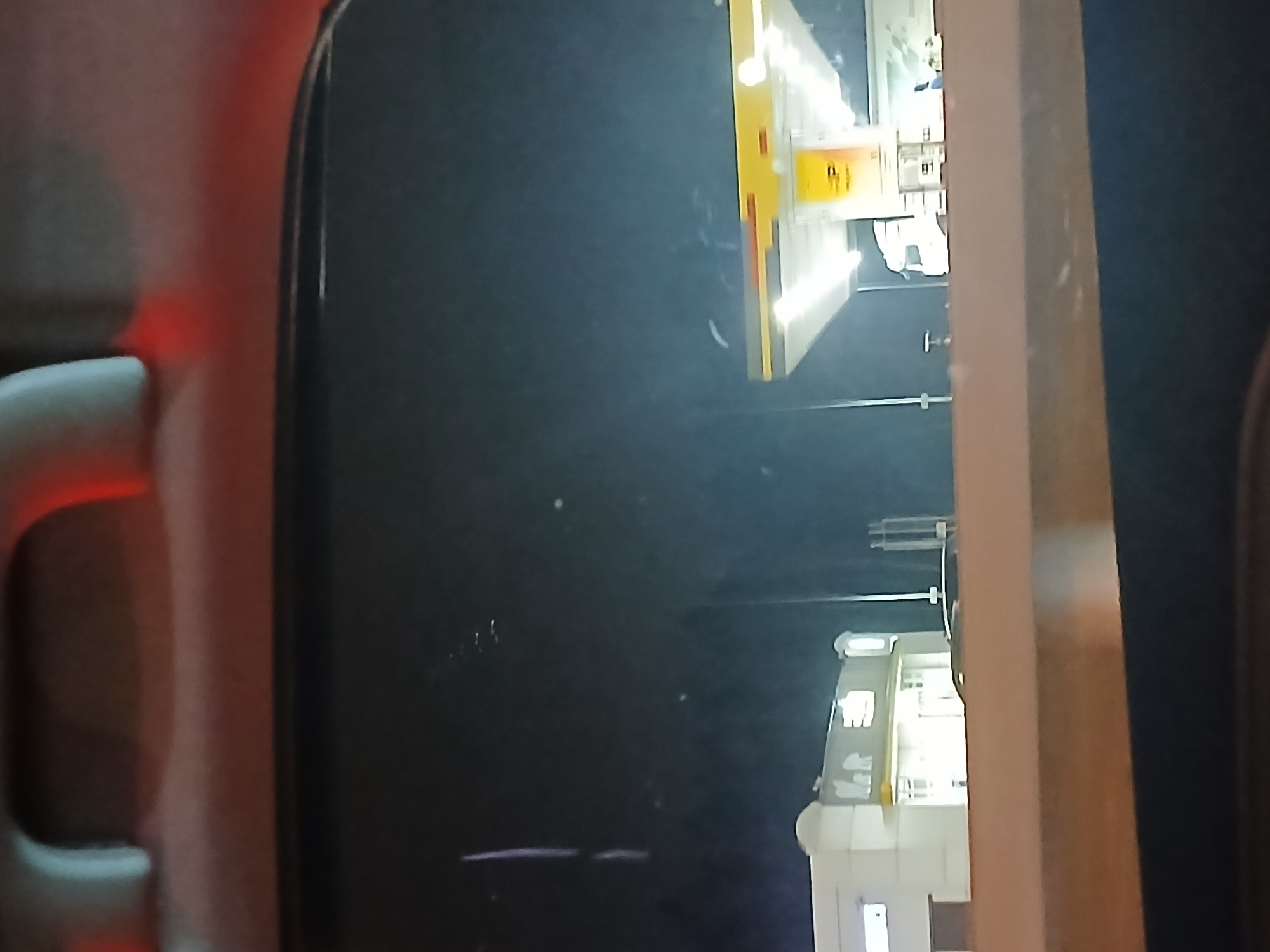 The image depicts an exterior night scene observed from within a vehicle, likely through a windshield or window, with a portion of the car's interior visible at the top (headliner and grab handle with red light reflections). The location is the city of Unknown, Unknown. The primary subjects are two commercial buildings, presumably part of a service station or truck stop. On the left, a multi-story white building with illuminated windows and a distinctive architectural style featuring arched elements and partially visible signage, including what appears to be "M R" and an "E." On the right, a brightly illuminated Shell gas station canopy, distinguished by its yellow and white design and the prominent yellow Shell Pecten logo on a black sign. Underneath this canopy, a large white semi-truck is parked. The environment is dark, indicating night, with significant artificial illumination emanating from both structures. No individuals are clearly discernible.