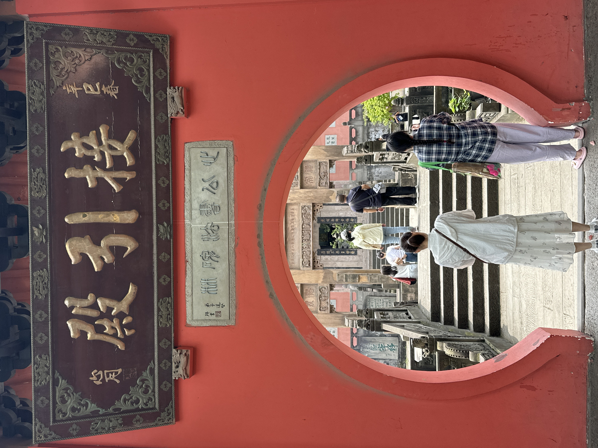 Captured in the Jiulongpo District of China, the image presents a vivid scene viewed through a circular red doorway. A dark brown plaque, adorned with Chinese characters in gold, hangs on the left side of the entrance. To the right, another smaller stone inscription displays similar calligraphy.
People populate the scene within the doorway. Two women stand close to the viewer, with one in a white dress and the other in a checkered shirt, walking away from the camera. A few people ascend the stone steps, and some are exploring the area further into the complex, revealing an array of traditional Chinese architecture. Buildings with carved stone details and inscriptions are visible in the background.