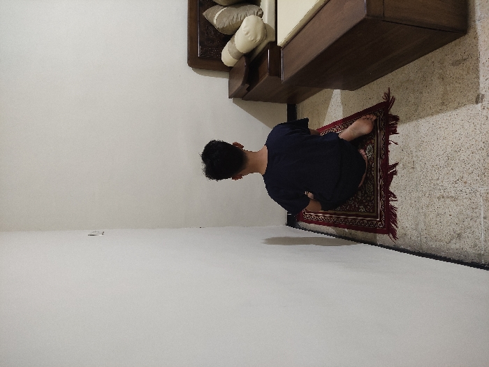 The image shows a person sitting on a prayer rug in the corner of a room. The person is wearing a dark blue t-shirt and has short black hair. The person is sitting with their legs crossed and their back straight. The rug is red with gold accents. The floor is tiled in a light beige color. There is a white wall to the person’s left and a wooden bed to the right. The bed has a white mattress and a brown wooden frame. There are two pillows on the bed. The room is lit by overhead lighting. The location, based on the coordinates, is in the city of Kecamatan Lawang, Indonesia. The coordinates provided place the location in a residential area of Lawang. The scene is set indoors with the person engaged in prayer.
