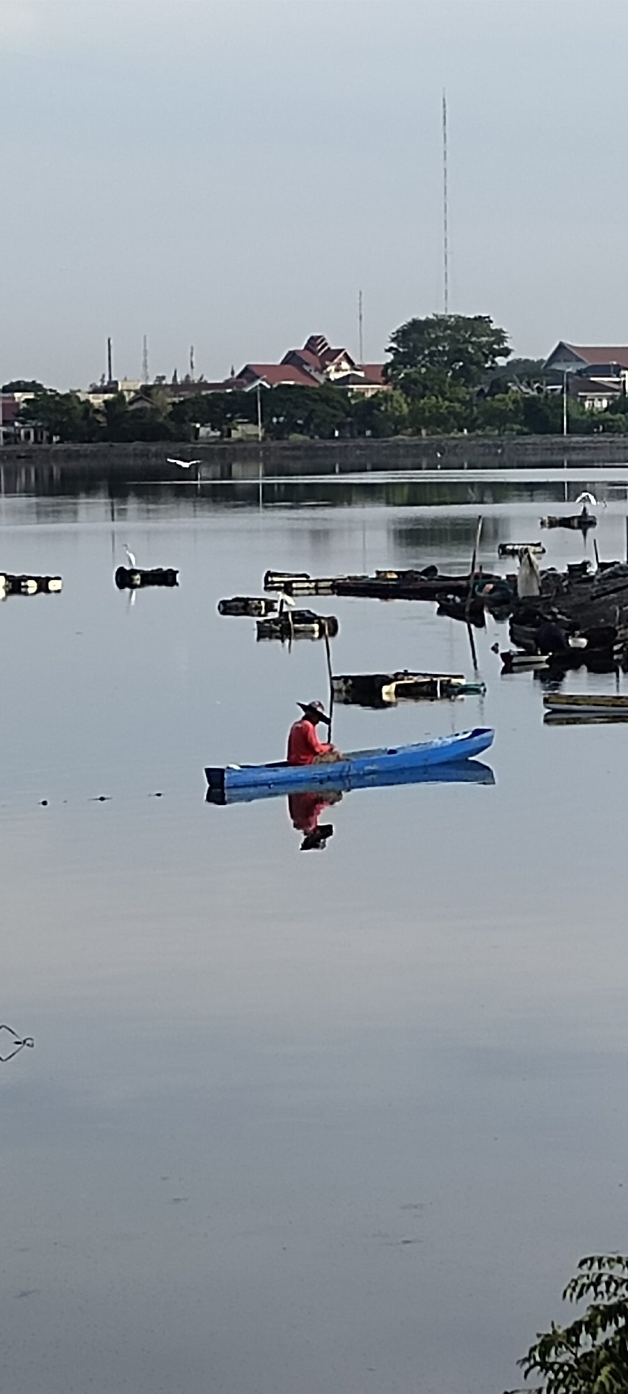 A single person in a blue kayak, wearing a red shirt and a wide-brimmed hat, is on a body of water. The kayak is angled slightly towards the right of the frame. The person's reflection is visible in the calm water below. In the background, a city skyline is visible with buildings and trees. There are also several floating structures in the water, some of which appear to be made of dark, rectangular objects. The sky is clear and pale. This scene is likely of the waters near the city of Lhokseumawe, Indonesia, possibly a bay or a river.