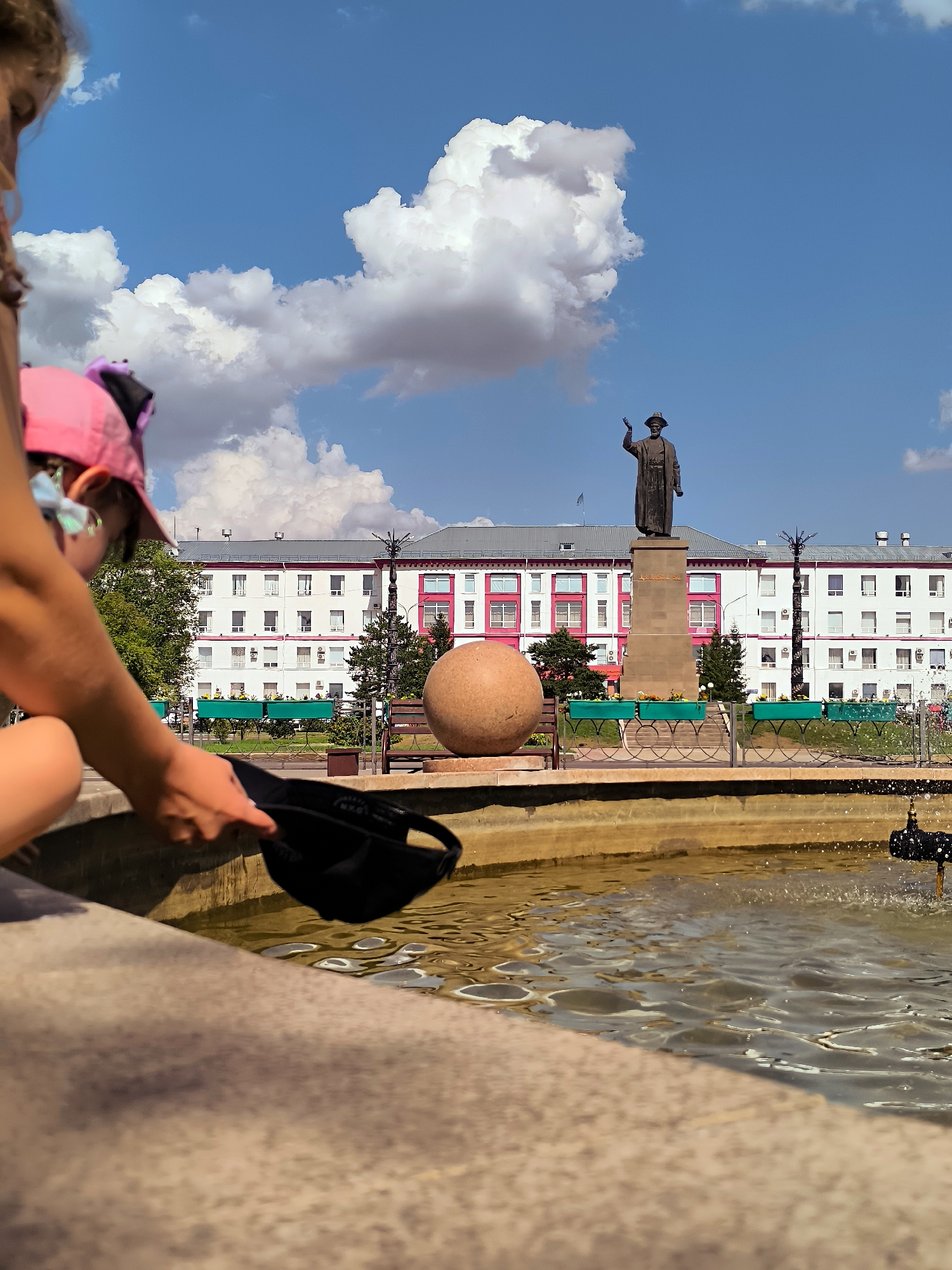 This is a medium shot of a park on a sunny day. In the foreground, the edge of a fountain and the hands of a person holding a black baseball cap are visible. The person is wearing a pink baseball cap and a light-colored top. In the midground, a large, smooth, spherical sculpture sits to the left of a bronze statue on a pedestal. The statue depicts a man in a hat and coat, with his right arm raised. Behind the sculpture and statue is a white building with red accents and many windows. Trees line the area in front of the building. The background is a bright blue sky with fluffy white clouds. This scene is likely located in the central park of Karaganda, Kazakhstan, given the presence of the Abai statue, a prominent landmark in the city.
