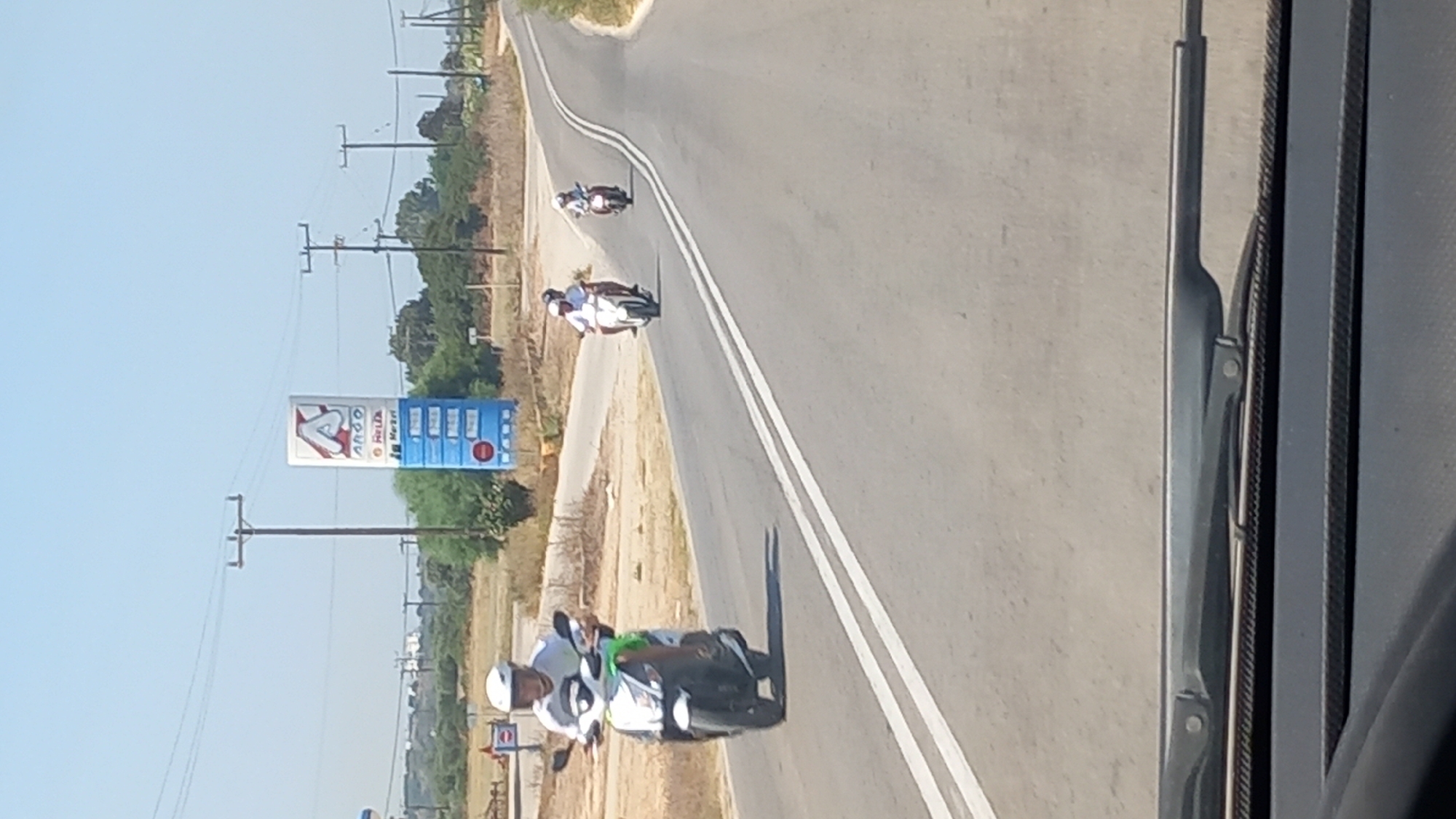 A clear, sunny day is depicted, with a light blue sky above. A road with white lines marking lanes is the primary focus. In the foreground, the edge of a car's dashboard and windshield wiper are visible, suggesting the photo was taken from inside a vehicle. On the road, several individuals are riding scooters.
The closest scooter is white and green, ridden by a person wearing a white helmet and shirt. Further down the road, another scooter carries two people, both wearing white. A third scooter, further in the distance, has a single rider.
To the left of the road, a tall, multi-colored signpost stands. It features a prominent red "A" logo at the top, followed by text that appears to be related to a gas station or convenience store, with smaller blue panels displaying what might be prices. Power lines and poles are visible crisscrossing the sky, and a few trees and dry vegetation dot the landscape. The overall impression is a rural or suburban setting on a bright day.