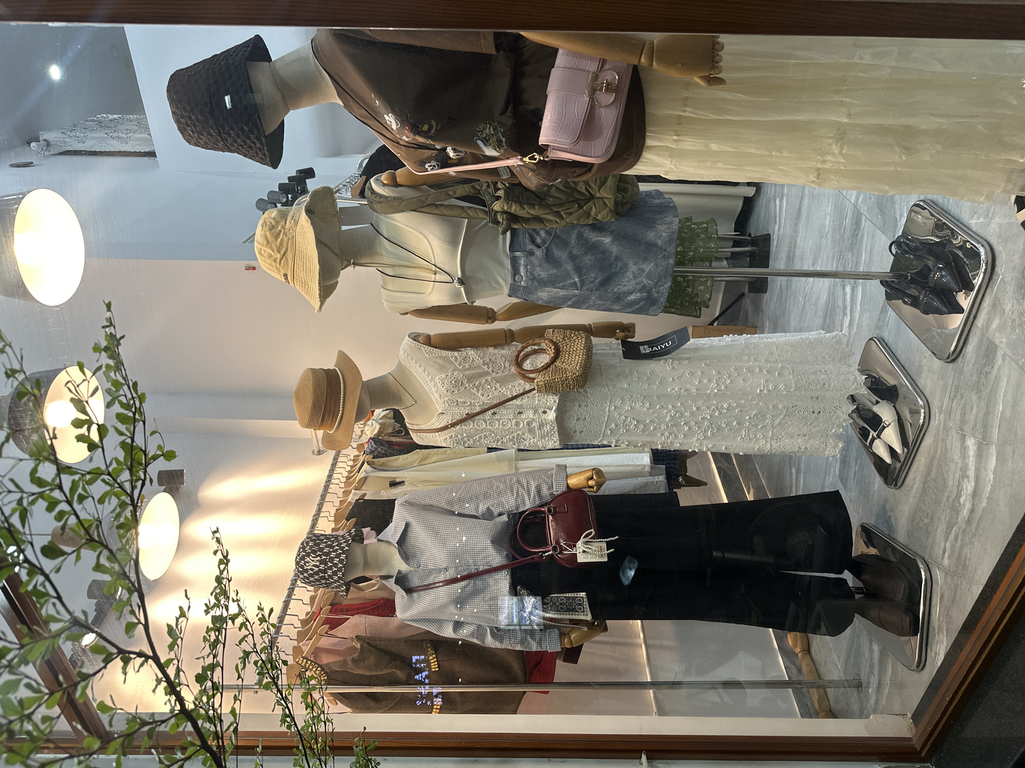 A retail storefront, likely a clothing boutique, is located in Bac Nha Trang Ward, Vietnam. Mannequins wearing clothes are arranged vertically inside a glass display case. There are four visible mannequins. The top mannequin is wearing a brown top, a light-colored skirt, and a dark-colored hat, and has a pink purse. The next mannequin wears a light-colored top, denim bottoms, and a hat. The third mannequin wears a white top and skirt and a hat. The bottom mannequin wears a striped shirt, dark pants, and a hat and has a burgundy purse. Clothes hang on a rack behind the mannequins. A plant with green leaves is visible in front of the storefront. Lights are reflecting in the glass. The floor is gray. Shoes are displayed by each mannequin.