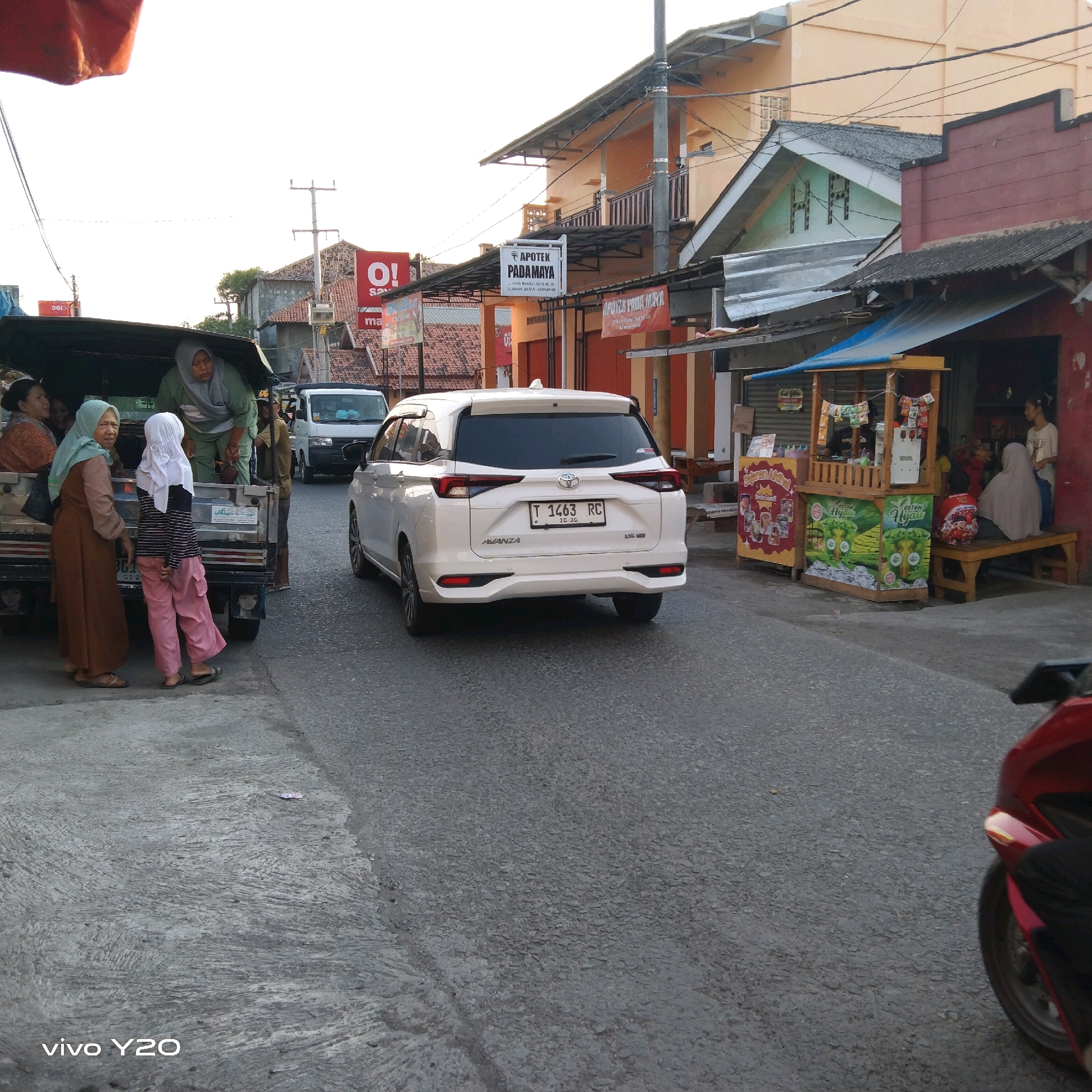 This is a street-level view in Karawang, Indonesia, capturing a busy market or roadside scene. In the foreground, a red motorcycle is partially visible on the right side of the frame. The road surface transitions from concrete on the left to asphalt on the right. Several people are gathered around the back of a pickup truck on the left, with some individuals appearing to be passengers and others potentially vendors. The truck itself has a dark canopy.
Further down the street, a white Toyota Avanza is driving away from the camera. On the right side of the road, a small roadside stall with a blue canopy is set up, selling drinks. Several people are seated on a wooden bench in front of the stall, and others are standing. Buildings line both sides of the street, displaying various signs, including one for "APOTEK PADAMAYA". The sky is bright, suggesting daylight, possibly late afternoon or early morning. The overall impression is of a vibrant, everyday scene in a commercial area.