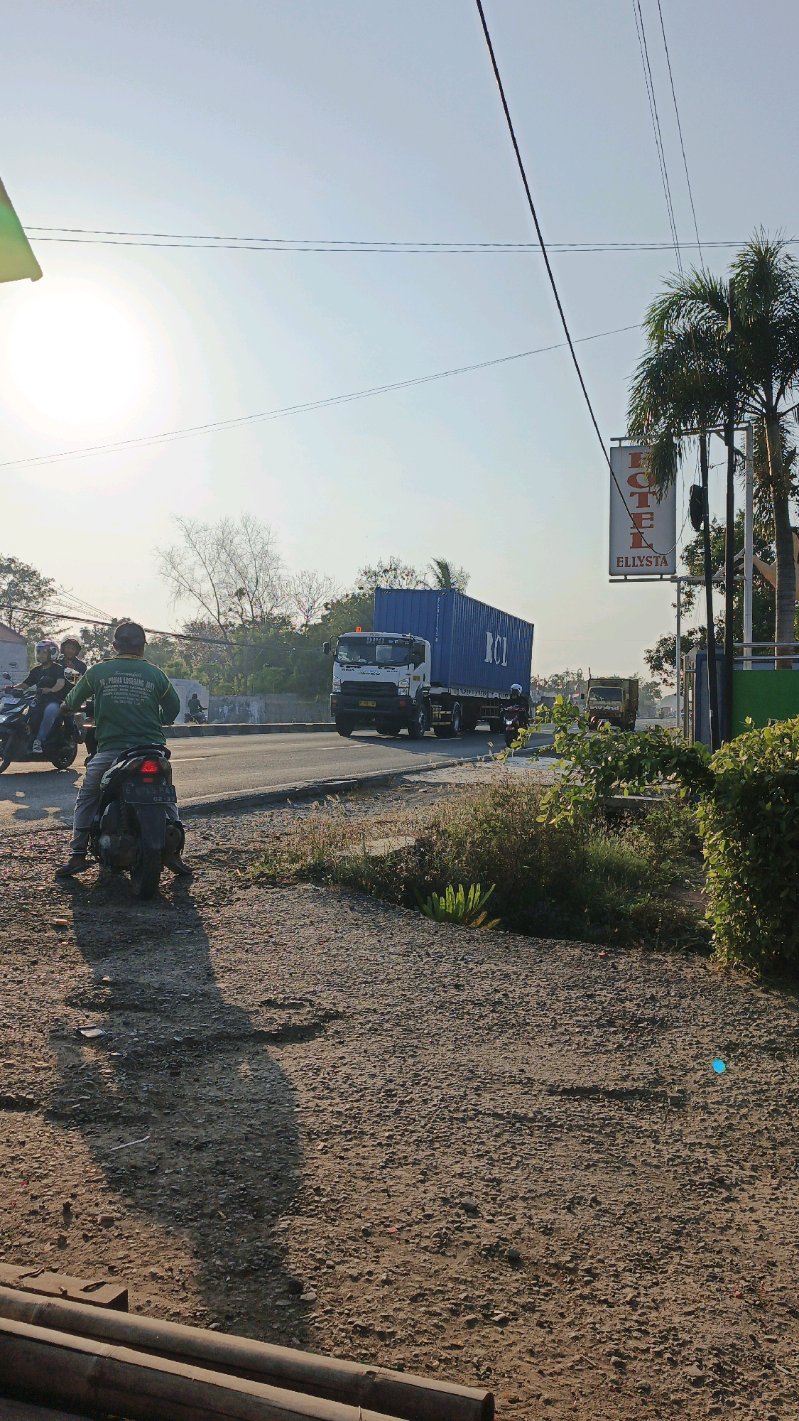 The image shows a view of a road in Krimun, Indonesia, during the daytime. The sun is bright and low in the sky, casting long shadows. A large container truck with "RCL" written on the side is driving on the road. There are several motorcycles, some with riders, also on the road or parked nearby. In the foreground, there is a dirt and gravel area with some logs. A sign for "HOTEL ELYSTA" is visible on the right side of the image. The environment appears to be semi-urban or suburban, with some trees and vegetation visible.