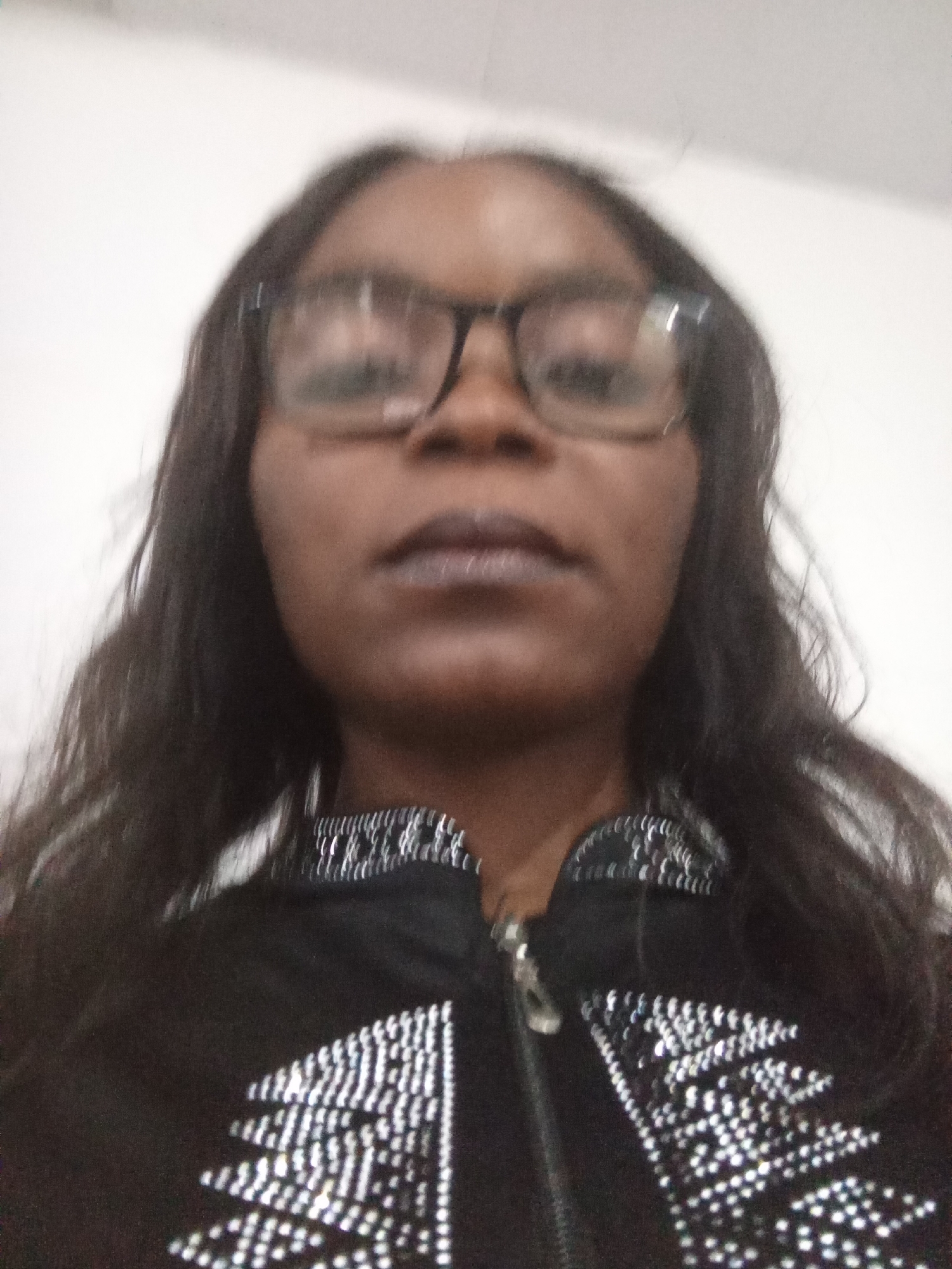 The image is a selfie of a woman wearing glasses and a black jacket with rhinestone embellishments. The woman has long, dark hair. She is looking directly at the camera. The background appears to be a plain wall, likely indoors. There is no indication of the city of Kason, Nigeria, or any specific urban environment in the image. The photo is slightly out of focus.