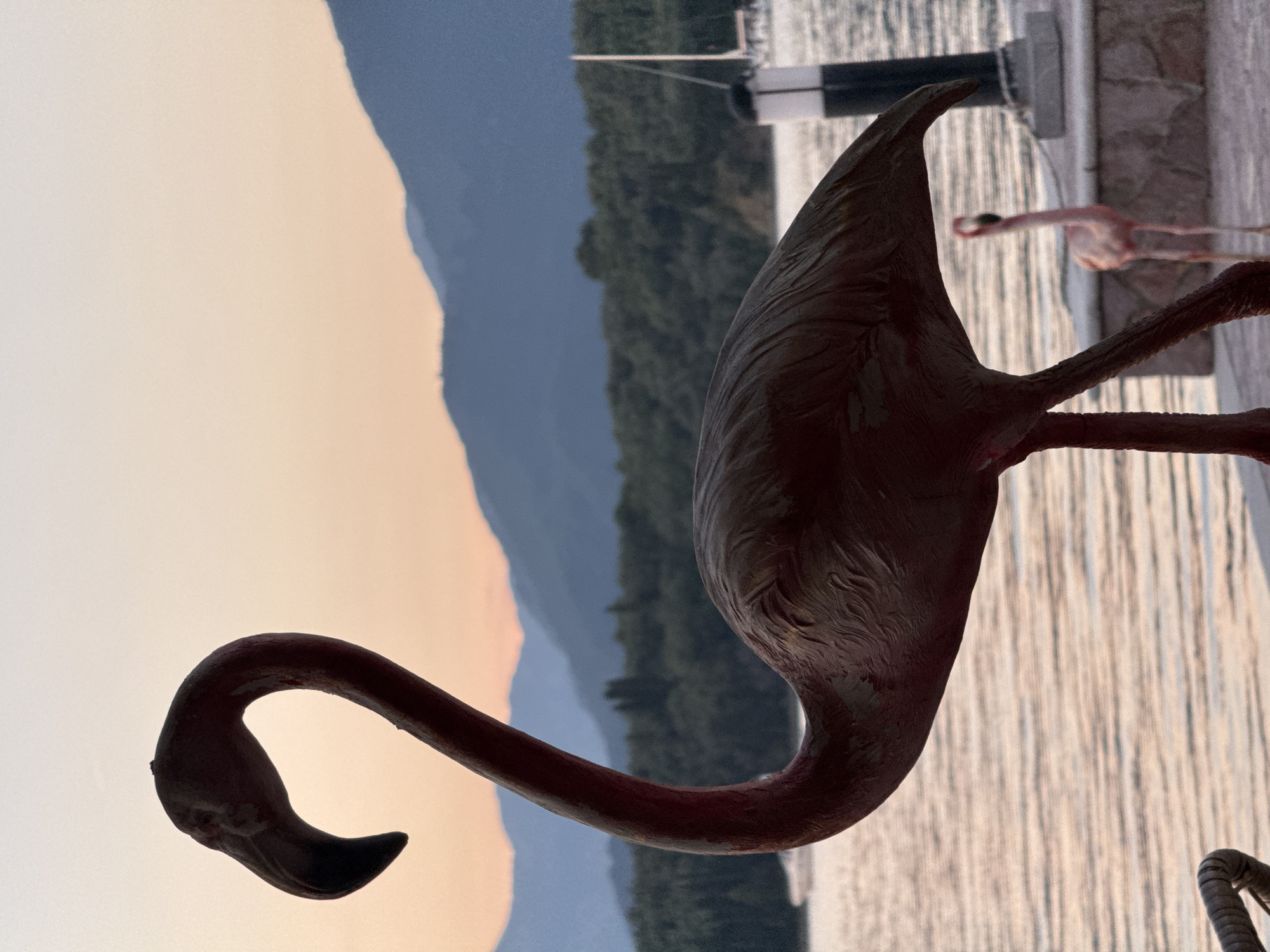A flamingo statue stands on a concrete waterfront in Bogišići, Montenegro. In the background, a lake and mountains are visible at dusk. Another flamingo statue is partially visible in the background on the right. A portion of a pier or dock with a dark piling is visible in the background on the right.