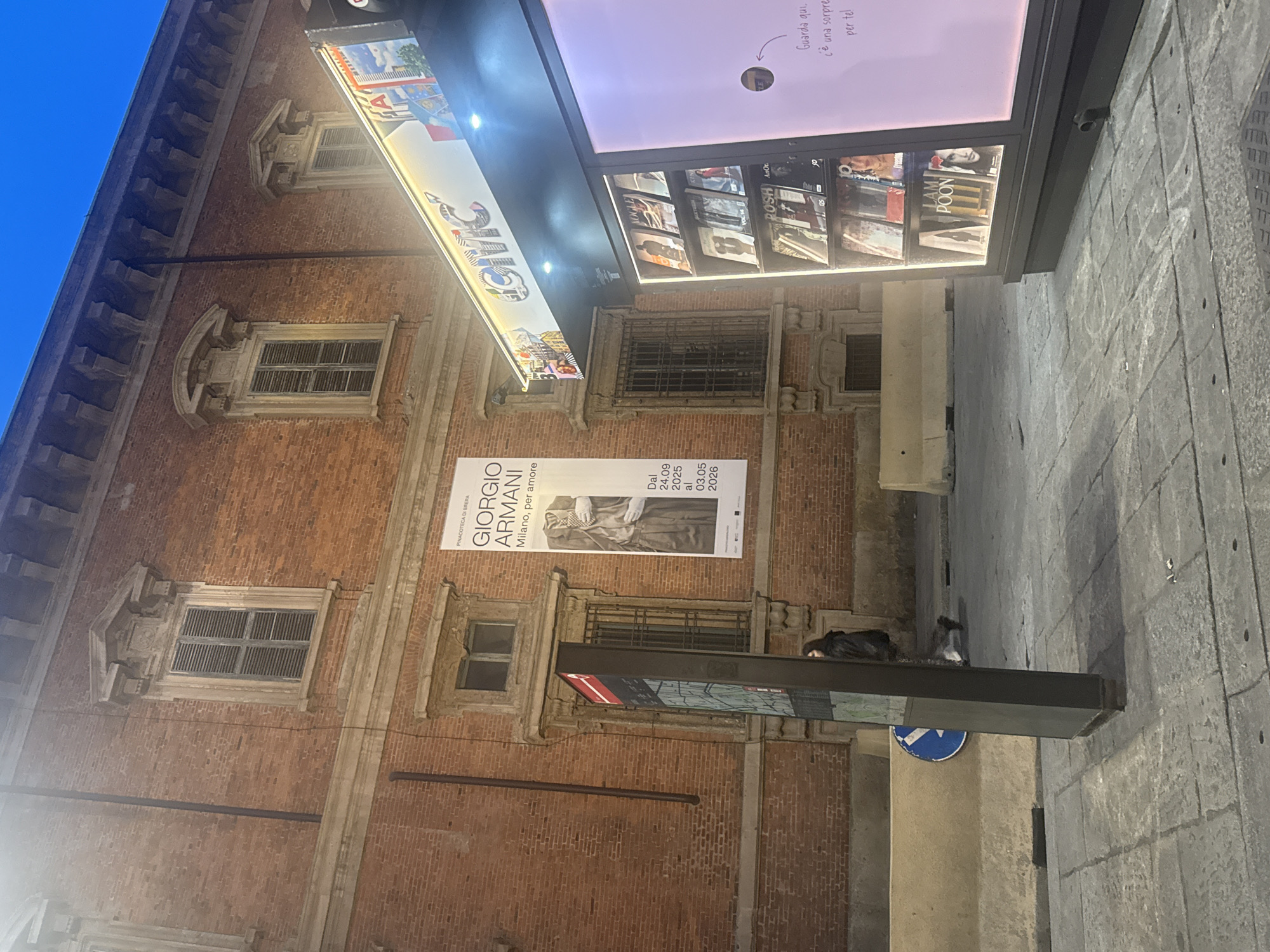 The image shows a street scene in Milan, Italy, at dusk or night. The main subject is the Palazzo di Brera, a historic brick building with ornate window frames and closed shutters, possibly housing the Pinacoteca di Brera art gallery.
A large promotional banner for a Giorgio Armani exhibition is attached to the building, displaying dates "24.09.2025 al 03.05.2026." To the right, the word "CIVIC" is visible, as well as illuminated displays featuring magazines and a pink area with text.
The sidewalk, paved with rectangular stones, has a street sign with a directional arrow and concrete structures, including a modern information display.
Two people are visible in the scene. One is standing and wearing dark clothing. The second person is lying down.