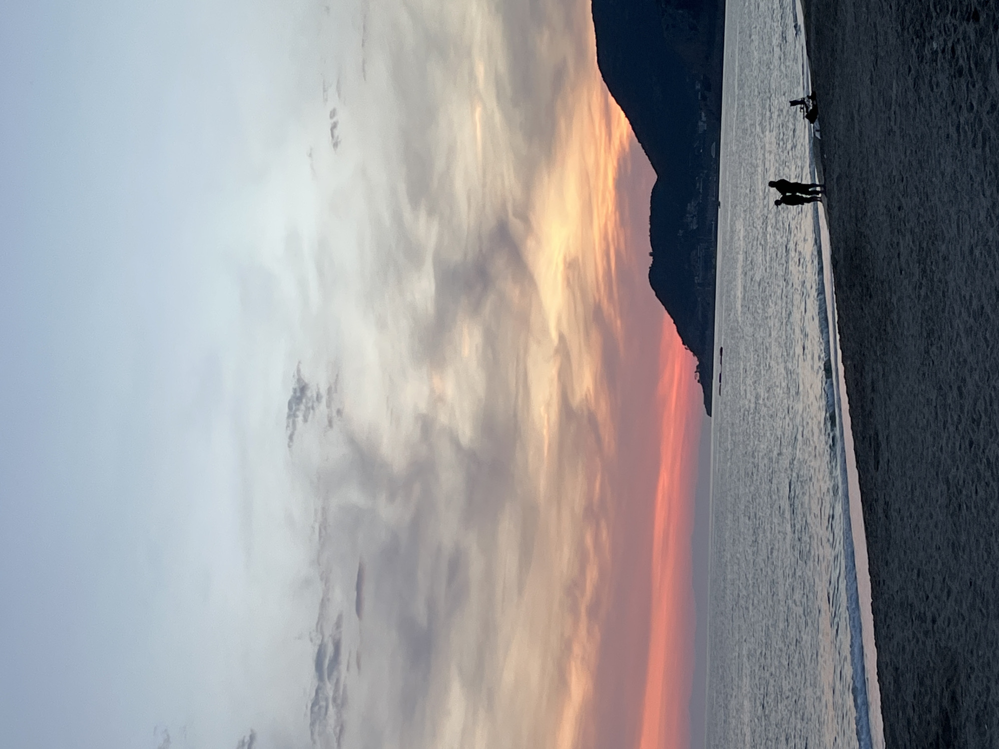 The image shows a beach scene in Alanya, Turkey at sunset. The sky is filled with dramatic clouds, transitioning from grey and white to orange and red near the horizon. To the right is a dark, silhouetted mountain. The sea reflects the colors of the sky. On the beach, the silhouettes of two people are visible near the water's edge, with another individual or object slightly further down the beach. The sand is dark.