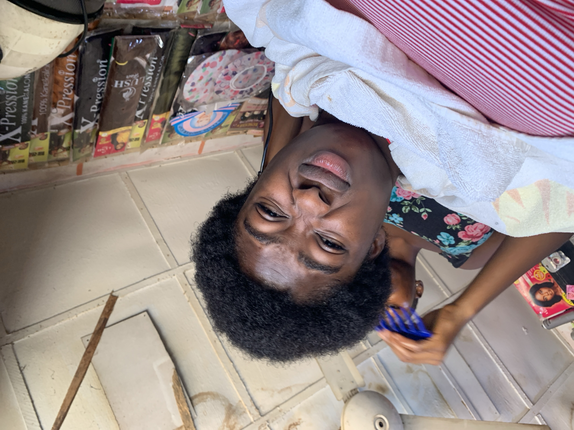 The image shows a person lying down and getting their hair combed. The person has dark skin and dark, curly hair. They are wearing a black tank top with a floral pattern. A white towel is draped over their chest and shoulder, and a red and white striped fabric is visible in the background. The person is holding a blue comb and appears to be looking at the camera with a slightly pained or grimacing expression.
In the background, on a shelf, are several packages of hair products. Some of the packaging has text that is partially visible, including "X-Pression" and "100% Kanekalon." There are also some colorful circular designs on some of the packages. The floor is tiled with a light-colored material. The overall setting appears to be indoors, possibly a salon or a home. The lighting suggests it is daytime.
The image was taken in Rafin Par, Nigeria.