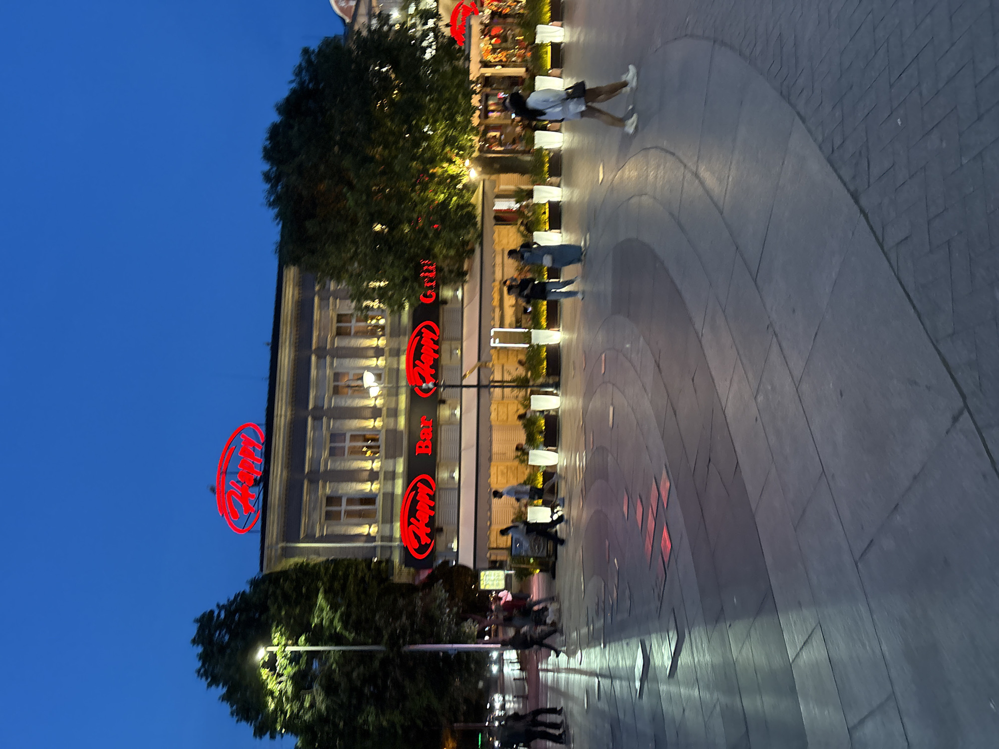 In Varna, Bulgaria, at dusk, a wide pedestrian walkway is illuminated by embedded lights and streetlights. In the background, a multi-story building features large red illuminated signage for "Happy Bar & Grill." Several trees line the front of the building, their leaves lit by nearby streetlights. A few individuals are walking on the walkway. One woman in a white top and shorts walks towards the foreground, and another couple, dressed in darker clothing, walks away from the camera, heading towards the restaurant. The walkway itself is paved with large stone slabs and curved sections, with some square red lights embedded in the stone. The sky is a deep blue, indicating late evening.