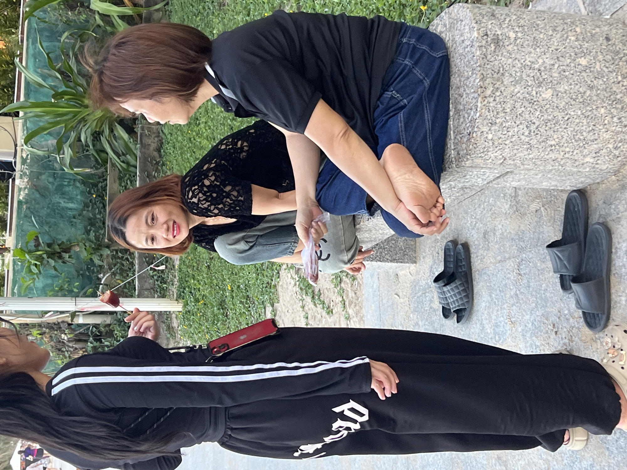 The scene depicts three women in an outdoor setting, likely a park or public space in Nha Trang, Vietnam, given the presence of tropical vegetation. Two of the women are sitting on a stone bench. One woman is wearing a black t-shirt and jeans, seated with her feet crossed on the bench. Adjacent to her sits another woman with auburn hair, dressed in a black lace top and green pants. A third woman is standing, and is wearing a black track suit with white stripes down the side, holding a stick with what appears to be a sausage on it. Footwear, including slides, is visible on the pavement near the bench.