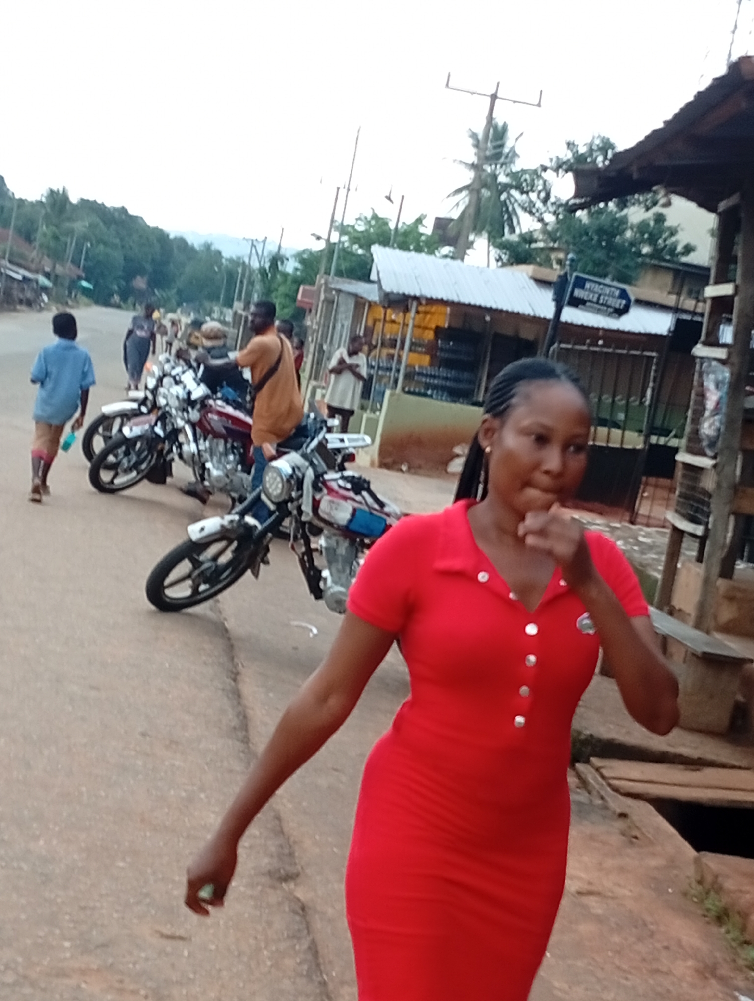 A medium shot captures a young Nigerian woman in a red dress walking on a dirt road in Nsugbe, Nigeria. She has dark braided hair and is looking forward with her hand near her mouth. Two motorcycles are parked on the side of the road, and several other people are visible in the background. The environment appears to be a rural or semi-urban area with buildings and vegetation. A street sign with "NYAONTVH NIWEKE STREET" is visible on the right side of the image.