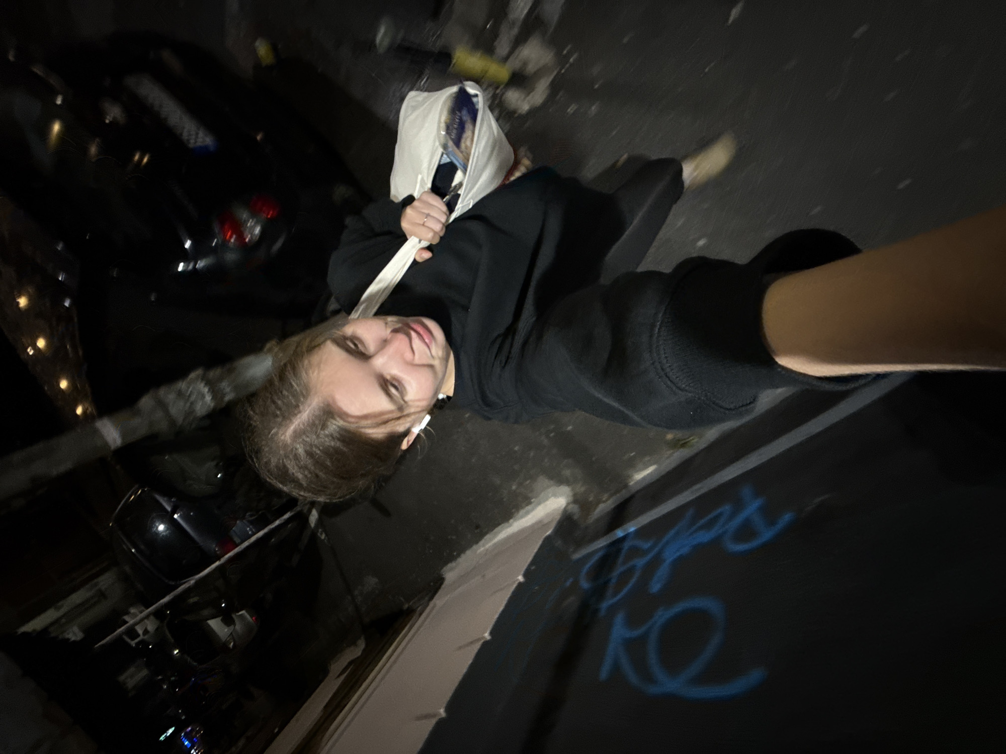 A low-angle, distorted perspective photograph taken at night. A person, presumably the photographer, is visible from the chest up, wearing a black hoodie with the sleeve pulled up to the elbow on the right arm. The person is also wearing what appears to be an earbud. They are holding a white tote bag with visible contents. Beside the person is a dark-colored surface with light blue graffiti on it. In the background, on the left side of the image, are multiple dark-colored cars parked along a street, and buildings with illuminated windows. The ground appears to be paved and wet. The location is likely an urban area in Belgrade, Serbia based on details like the type of vehicles, architecture, and street layout.
