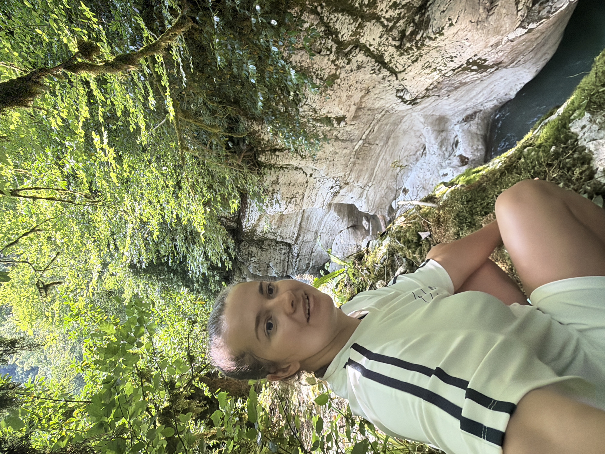 In Khostinskiy, Russia, a woman with her hair tied back is seated on a moss-covered ledge overlooking a narrow, clear river flowing through a deep, limestone gorge. The gorge walls are sheer and sculpted by erosion, with distinct horizontal striations. Lush green foliage and trees, some draped with moss, overhang the gorge, creating a shaded and natural environment. The woman is wearing a white t-shirt with black stripes on the sleeves and white shorts. She is looking to her right, with a slight smile. The sunlight filters through the leaves, illuminating parts of the scene and creating shadows.