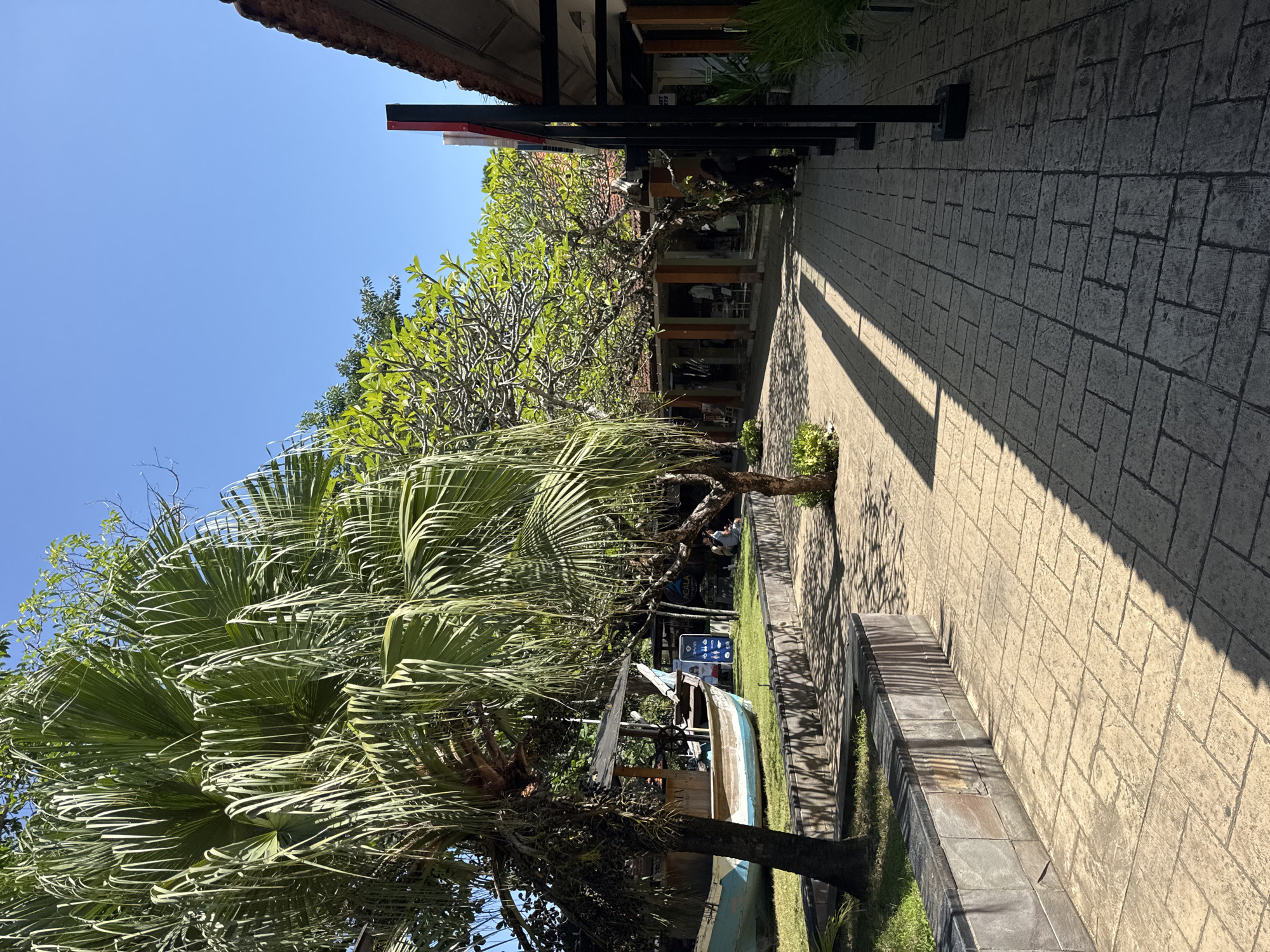In Kuta Selatan, Indonesia, a paved walkway stretches through a tropical outdoor area. To the left, a large palm tree stands prominently, its fronds casting shadows on the ground. Adjacent to the palm tree is a weathered, light blue and white boat, partially covered by a wooden structure with a thatched roof. A low, stone retaining wall runs along the edge of the walkway, separating it from a grassy area.
The walkway is constructed of rectangular, beige paving stones arranged in a staggered pattern. Sunlight casts distinct, linear shadows across the stones, indicating a bright, sunny day.
In the background, a row of buildings with terracotta tile roofs lines the right side of the walkway. These buildings appear to be shops or establishments, with open-fronted entrances revealing glimpses of interiors and displays. Several trees with lush green foliage are interspersed among the buildings, providing shade and a natural element to the scene.
A few people are visible in the distance, casually walking or sitting near the establishments. One individual is seen standing near the entrance of a building on the right. The overall environment suggests a relaxed, resort-like setting, typical of coastal tourist destinations.