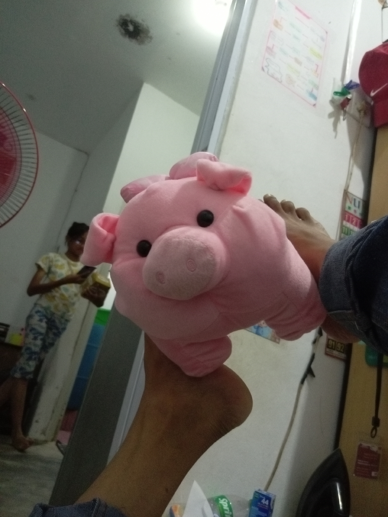 A close-up, low-angle shot focuses on a pink pig plush toy held by a person's arm and foot, positioned in the foreground. The plush toy is soft, with black button eyes and floppy ears. To the left and slightly behind the pig, a young person wearing a patterned t-shirt and camouflage-patterned pants is visible, holding a phone and a yellow object. The background reveals an indoor setting with a white wall and a partial view of a red-bladed fan on the far left. A faint smudge is visible on the ceiling. The overall lighting is dim, with a light source seemingly coming from the right. The scene appears to be a casual indoor environment, possibly a residential area in Batam, Indonesia.