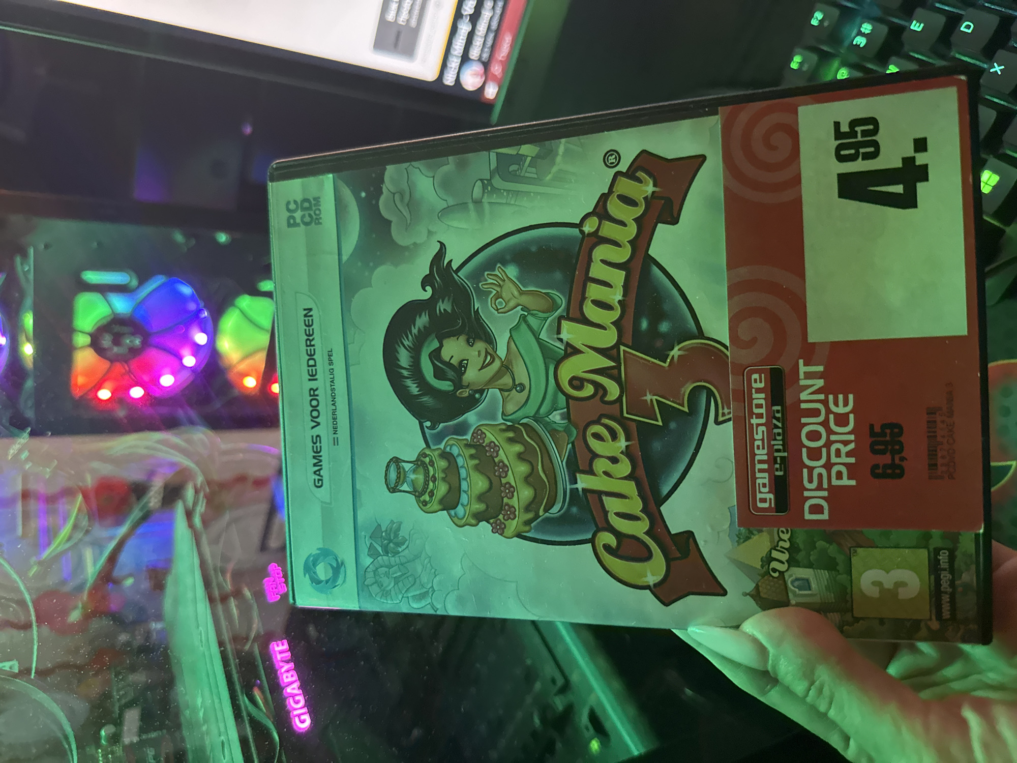 The image shows a close-up of a hand holding a PC game case for "Cake Mania 3". The game case has a discount sticker on it indicating a price of "6,95" and another sticker indicating a discounted price of "4,95". The background is out of focus and appears to be a computer setup with RGB lighting. The image does not provide any information about the city of Pazardzhik, Bulgaria, or any other geographical location.