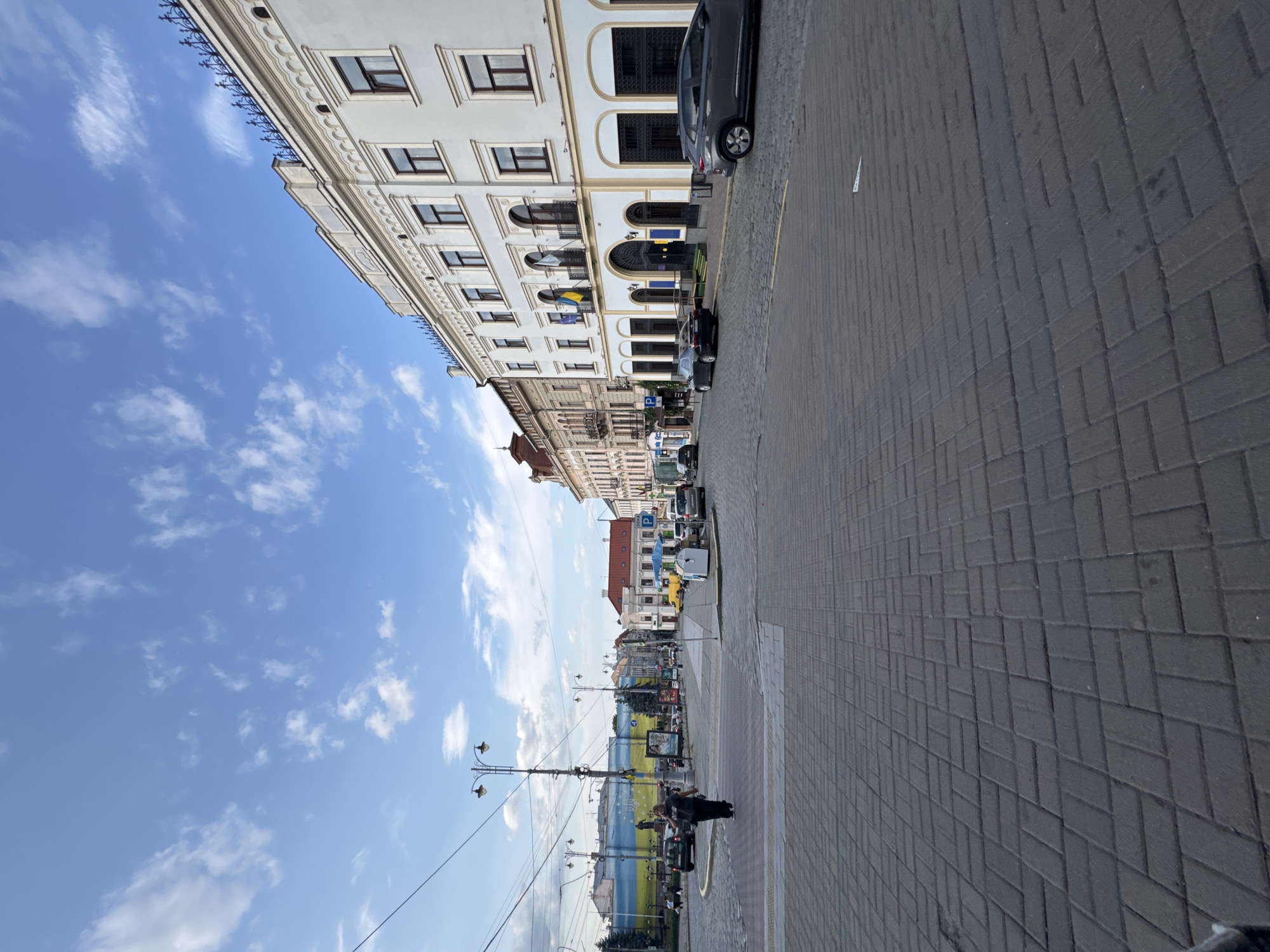 The image depicts a street scene in Chernivtsi, Ukraine. The perspective is from a slightly elevated position looking down a cobblestone street lined with buildings. In the foreground, the cobblestone pavement is clearly visible, with a few diagonal white lines indicating pedestrian crossings or road markings.
To the right, a modern white building with arched ground-floor windows and a decorative cornice dominates the view. Ukrainian and European Union flags are displayed on the facade of this building. Several cars are parked along the street in front of this building, including a dark grey SUV and a dark sedan.
Further down the street, on the left side, a large mural depicting the Ukrainian flag and national symbols is visible on a building. More traditional European-style buildings with multiple stories and balconies line the street, showing varying architectural styles and roof colors.
Several cars are in motion or parked in the middle ground of the scene. A woman in dark clothing is walking across a pedestrian crossing in the mid-left portion of the image. Streetlights and utility poles are present throughout the scene, with overhead wires crisscrossing the sky. The sky is blue with scattered white clouds, indicating fair weather. The overall impression is of a bustling city street on a bright day.