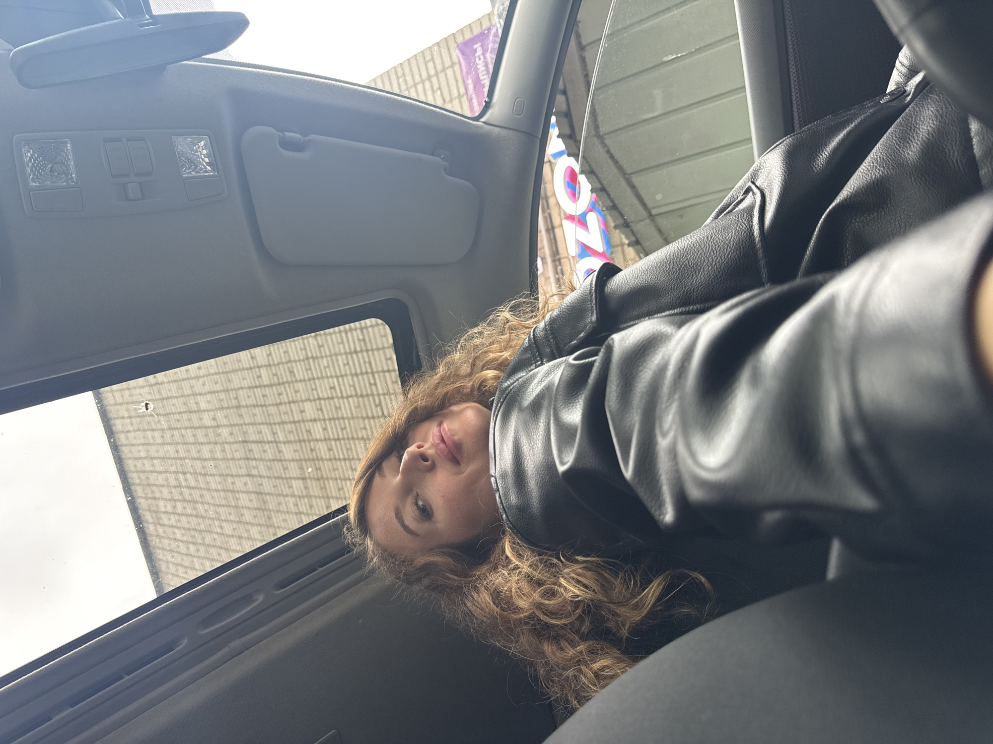 A woman with light brown, curly hair and a black leather jacket is captured in a low-angle selfie from within a vehicle. She is seated in the front passenger seat, looking upwards and directly towards the camera, with a slight smile on her lips. The vehicle's interior features a light grey overhead console with two rectangular dome lights and a central panel containing two rectangular buttons. A matching grey sun visor is visible on the passenger side. An open sunroof reveals a light grey sky and the upper portion of an exterior building facade, characterized by a repeating grid pattern, possibly concrete blocks or bricks. A small, dark, circular object or mark is present on the sunroof glass. Through the car's right-side window, exterior elements include parts of a building and fragmented signage. The letters "OZO" in blue and pink, indicative of the "OZON" brand, are visible on a sign. Above this, a purple banner displays the Cyrillic text "ЛИСЬ!" (likely part of "ЗАПИСЬ!", meaning "REGISTER!" or "APPOINTMENT!"). The scene is located in the city of Chaikovsky, Russia.