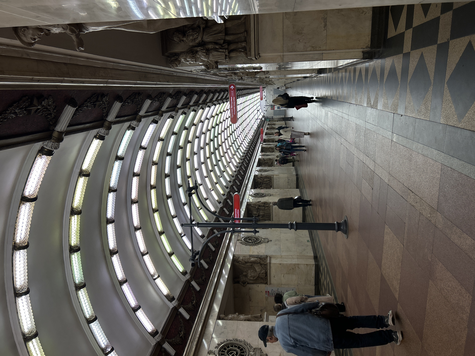 The image depicts a section of a metro station platform, presumably in Saint Petersburg, Russia, based on the architectural style. The station features a curved ceiling with multiple rows of illuminated light fixtures. On the left, the walls are decorated with classical architectural elements, including sculptures and ornamentation. Several people are seen walking along the platform or standing. A red directional sign is visible hanging from the ceiling. The floor is patterned with contrasting colored tiles.