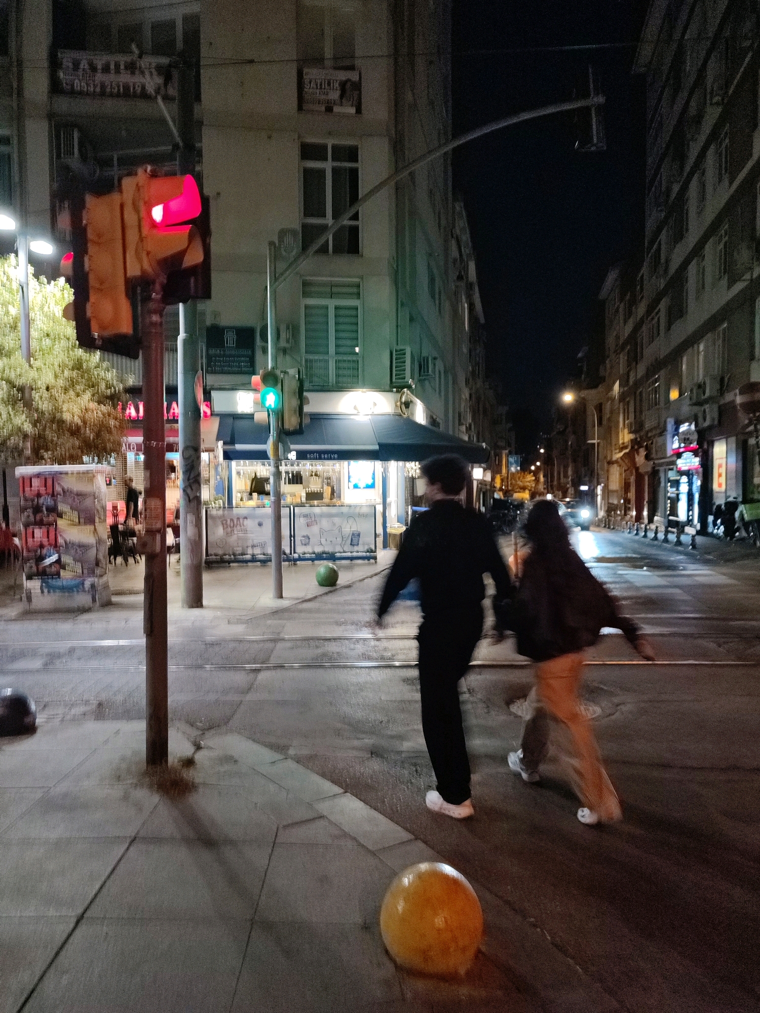 An image captures a street scene at night in Kadıköy, Turkey. A traffic light displays a red signal. Two figures, a person in a black jacket and another in a dark jacket with a person wearing light-colored pants, walk hand-in-hand across the street, their motion blurred. In the background, buildings line the street, some with lit windows and advertisements. A green traffic light is visible further down the street. The pavement is wet, suggesting recent rain. Various street elements are present, including a lamppost, bollards, and what appear to be tram tracks.