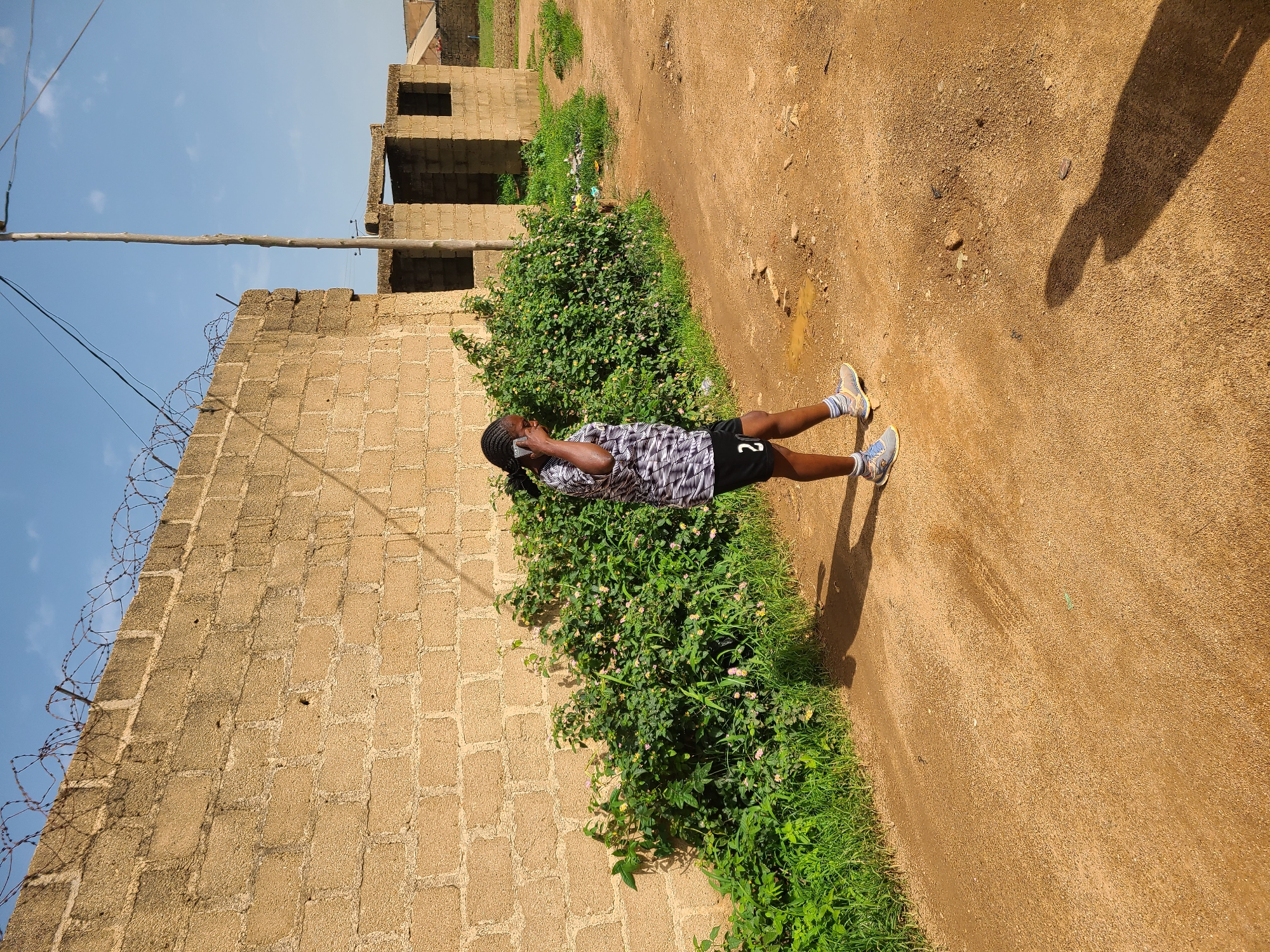 A young Black female with braided hair, wearing a black and white patterned t-shirt, black shorts with the number "2" on the left leg, and blue and grey athletic shoes with white socks, walks on a dirt path in Jos, Nigeria. She is holding a mobile phone to her left ear, appearing to be in conversation. Behind her, a rough, light brown brick wall topped with barbed wire runs horizontally across the frame. Adjacent to the wall is a dense patch of green foliage with scattered small pink and yellow flowers. Further in the background, unfinished brick structures with open doorways and window frames stand under a bright blue sky with scattered white clouds. The ground is a reddish-brown dirt, marked with puddles of water and scattered pebbles. A wooden utility pole with wires extends vertically towards the sky. The lighting suggests it is daytime, with shadows cast by the woman and the wall.