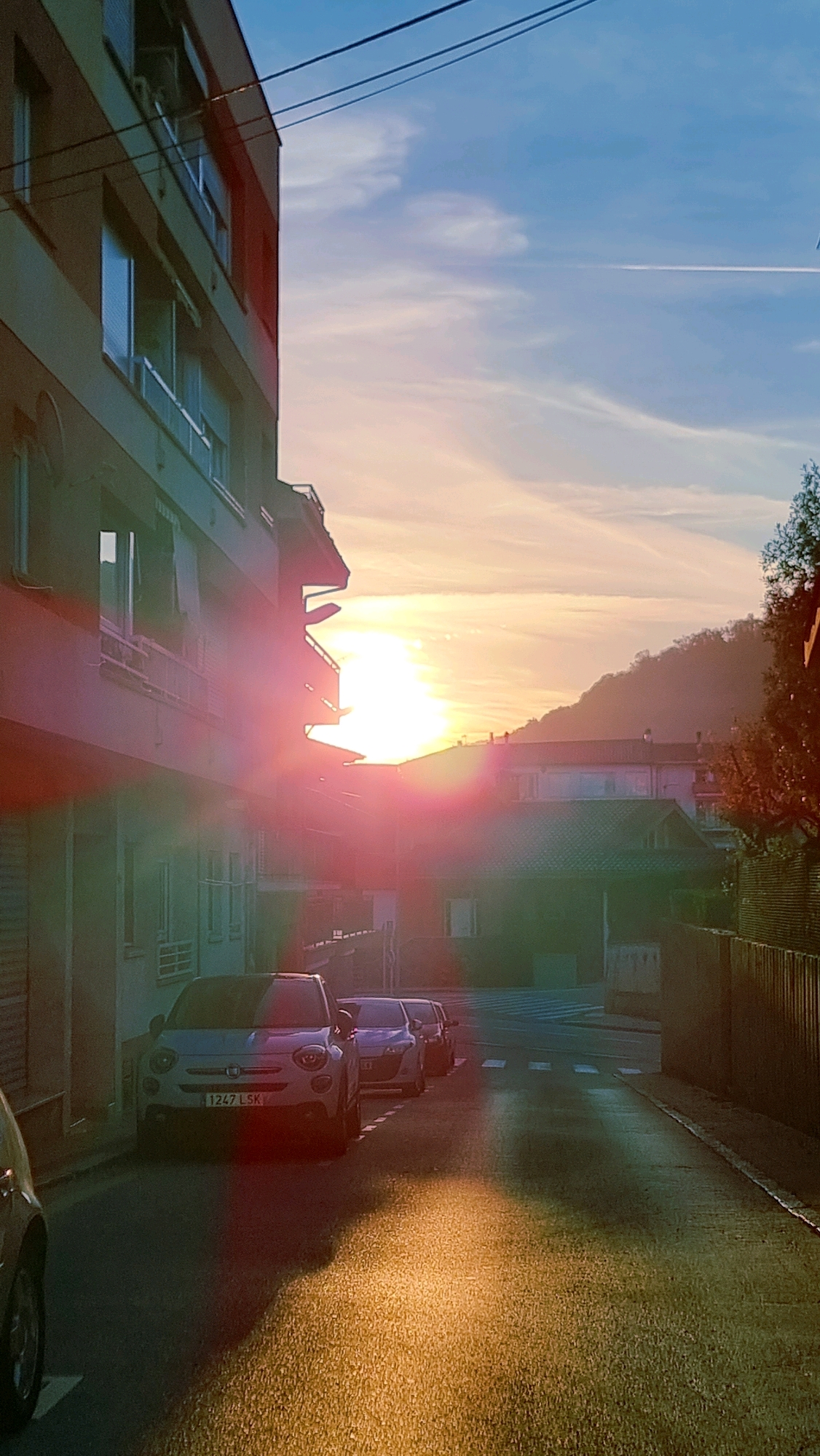 The image depicts a street in Sant Joan Les Fonts, Spain, during sunset. A multi-story building with balconies and windows lines the left side of the street. Several cars are parked along the right side of the road, including a white Fiat 500 with the license plate "1247 LSK". The setting sun casts a warm, golden glow on the asphalt and illuminates the sky with shades of orange and pink, with a contrail visible from an airplane. A tree with autumn foliage is visible on the far right. The scene captures a quiet, residential street bathed in the evening light.