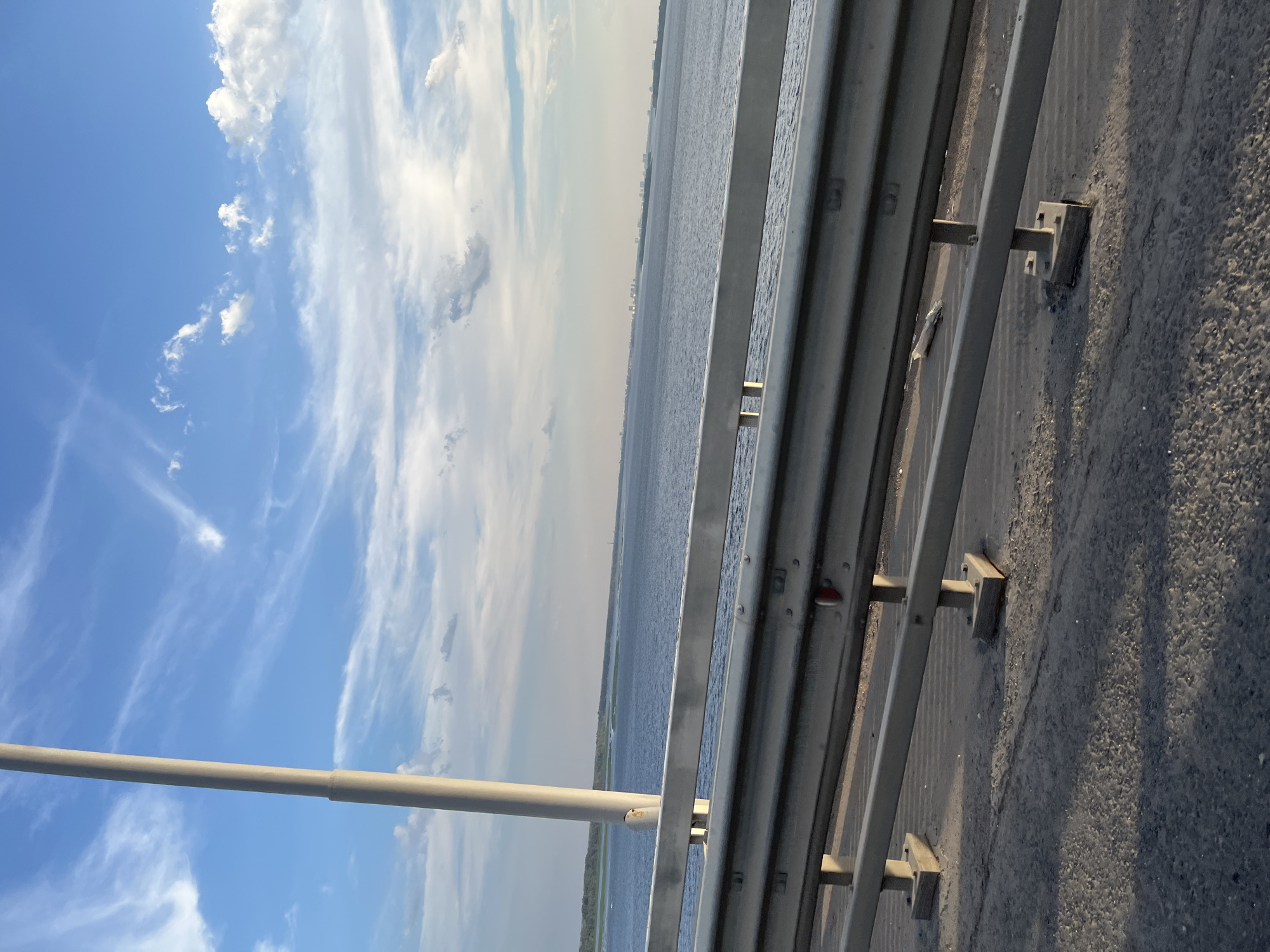 The image depicts a view from a bridge, looking out over a large body of water towards a distant cityscape under a partly cloudy sky. A tall, light-colored pole is visible on the left side of the frame, extending upwards towards the sky. A metal guardrail runs along the side of the bridge in the foreground, with concrete supports visible beneath it. The water is calm, and a faint line of green trees can be seen along the opposite shore. In the distance, a cluster of buildings suggests an urban area. The scene appears to be set in Saratov, Russia, likely on the Saratov Bridge crossing the Volga River. No people are visible in the image.