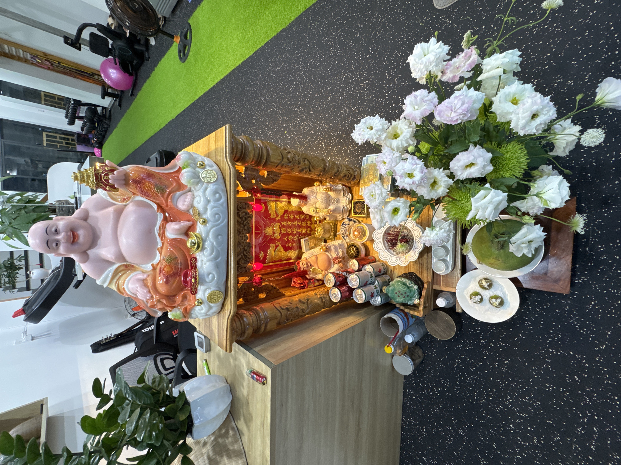 The image depicts an indoor scene that appears to be a home or a private fitness space, likely located in Nam Nha Trang Ward, Vietnam. In the foreground, a wooden altar is set up with various religious figures and offerings. A large statue of a laughing Buddha is prominent on top of the altar, adorned with gold accents and sitting on a cloud-like base. Below this, smaller statues of deities and a frog figurine are visible, along with cans of beverages and other items. In front of the altar, a large bouquet of white and pink flowers is arranged, with a green fruit and several small white balls on a plate nearby. The background shows a gym or fitness area with exercise equipment, including treadmills, weight racks, and a bright green artificial turf floor. The lighting suggests an indoor environment, possibly with artificial lights illuminating the scene.
