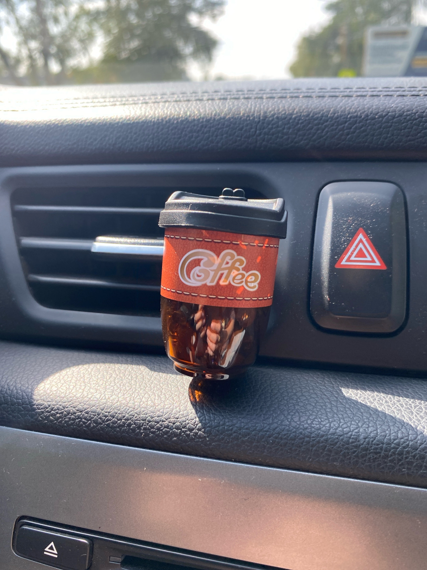 A car air freshener shaped like a miniature coffee cup is clipped to a car's air vent. The coffee cup air freshener has a brown liquid inside and a sleeve that says "Coffee". It is positioned to the left of the car's hazard light button. The car's interior is visible, with leather-like textured surfaces and a dark grey dashboard. Outside the car, out-of-focus greenery and sunlight suggest it is daytime. The location is the city of Kudinovo, Russia.