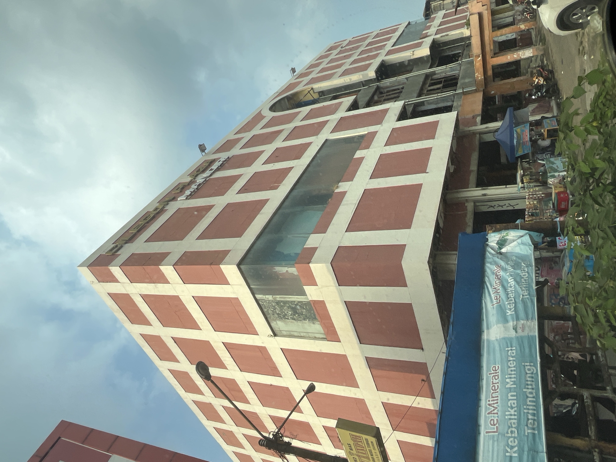 The image depicts a multi-story building with a distinctive facade of alternating red and white squares. The building appears to be a commercial establishment, possibly a mall or department store, given the visible signage. One sign, partially obscured by a banner, reads "Toko Mas Indah" which translates to "Beautiful Gold Shop". Another banner across the lower foreground, displaying "Le Minerale" and Indonesian text, suggests it is a distributor or retailer of bottled water.
The building is situated in Cilegon, Indonesia, as indicated by the prominent "Le Minerale" branding, a popular Indonesian bottled water brand. The street in front of the building is paved and appears to be a public thoroughfare. Several motorcycles are parked on the side of the street, and some small stalls or kiosks are set up near the entrance of the building. The sky is partly cloudy. The overall impression is that of a busy commercial area in an Indonesian city.