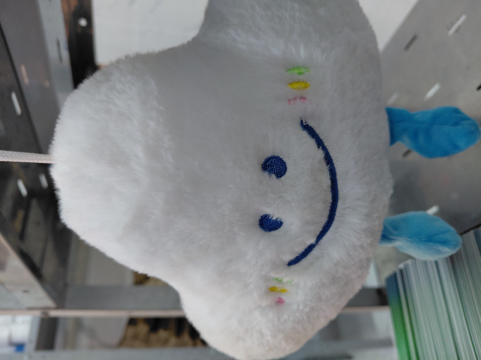 A close-up, eye-level shot captures a white, plush cloud-shaped toy with a smiling face embroidered in dark blue. The toy has two small, blue stuffed feet dangling below. On the left and right sides of the toy's face, there are clusters of tiny embroidered dots in pink, yellow, and green. The background is blurred but appears to be an indoor environment with metallic shelving and some stacks of colorful items, possibly paper samples, in the lower left corner. The lighting is even and diffused. The location is identified as Aktau, Kazakhstan.