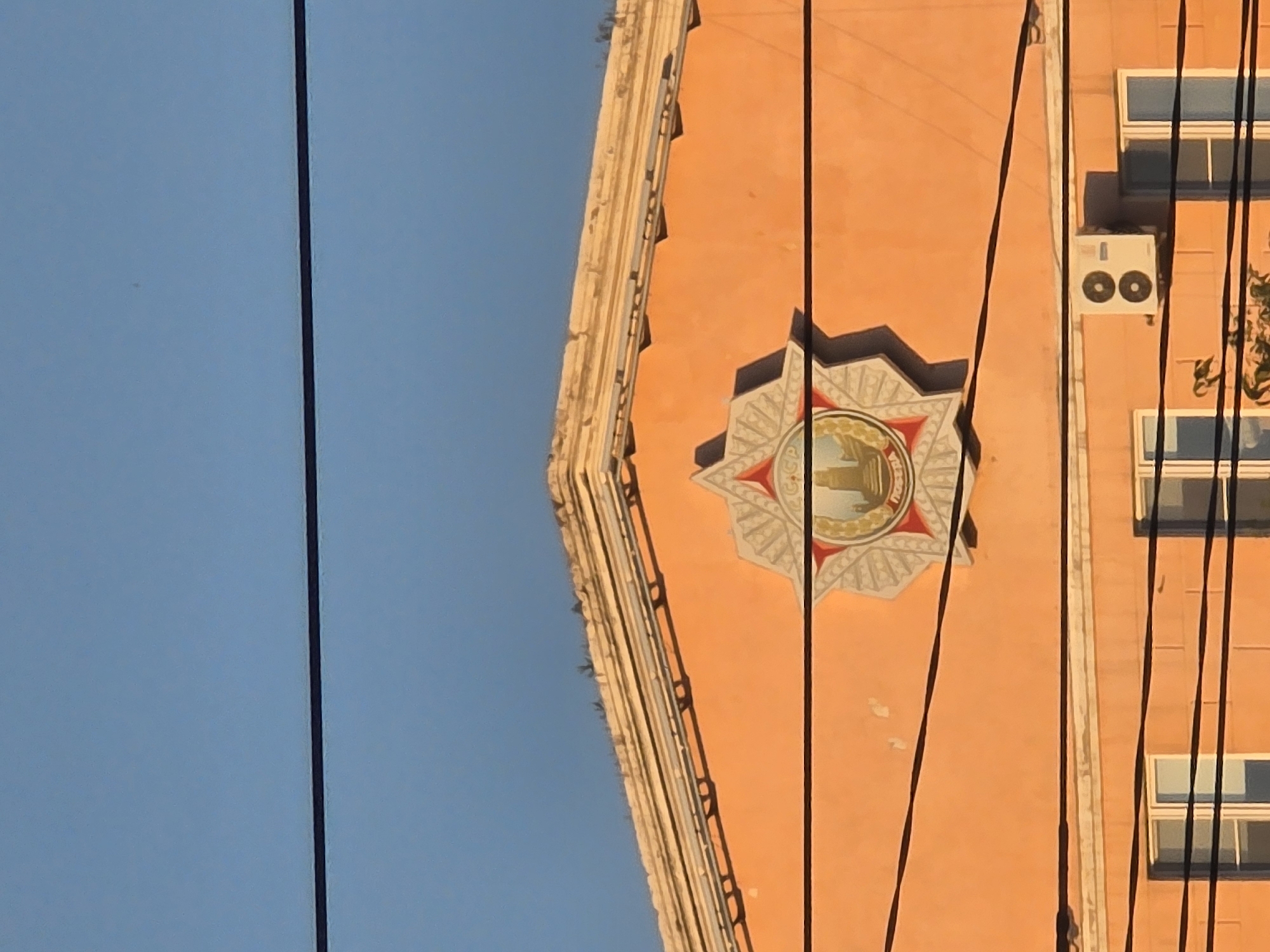 The image shows the facade of an orange building against a clear blue sky. The top of the building features a decorative cornice with layered trim. Centered on the facade is a circular emblem with an eight-pointed star shape overlaying it. Within the emblem, there is an image of a tower or monument and text. Below the emblem, a white air conditioning unit is mounted on the wall. The building has several windows with white frames. Power lines are strung horizontally across the image, obscuring parts of the building. The scene appears to be outdoors in an urban setting. This emblem is likely the coat of arms of the Republic of Buryatia, which features a Soyombo symbol and a ger, and is associated with the city of Ulan-Ude, Russia.