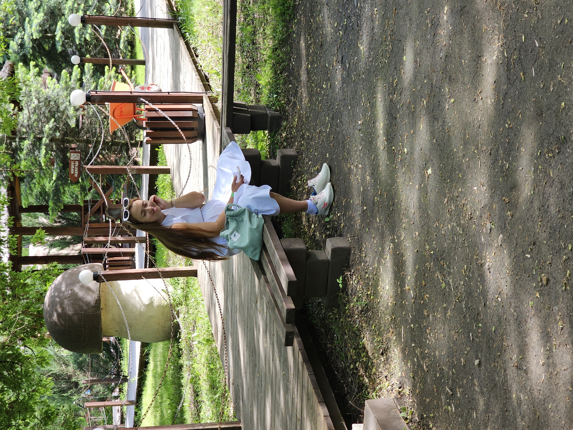 A young woman sits on a bench along a wooden walkway in Khimki, Russia. She is wearing a white dress and white sneakers with pink accents. A light green purse rests on the bench beside her. The walkway is lined with decorative elements, including large mushroom-shaped objects and wooden structures connected by chains. The ground to the right of the walkway is covered in dirt and fallen leaves. Vegetation is visible throughout the scene, suggesting a park or garden setting.